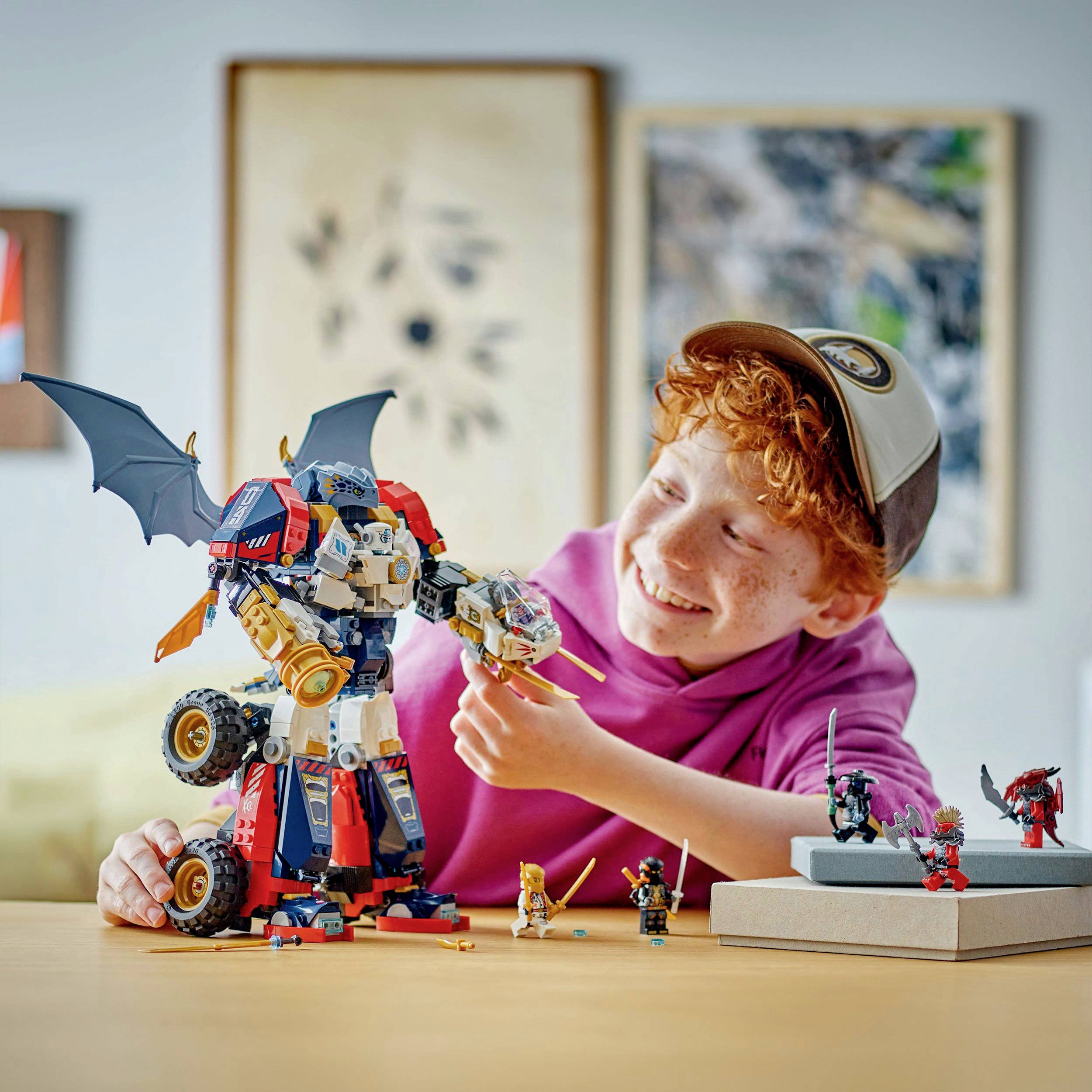 Een lachend kind met rood haar speelt met een grote, transformerende speelgoedrobot en kleine poppetjes op een tafel.