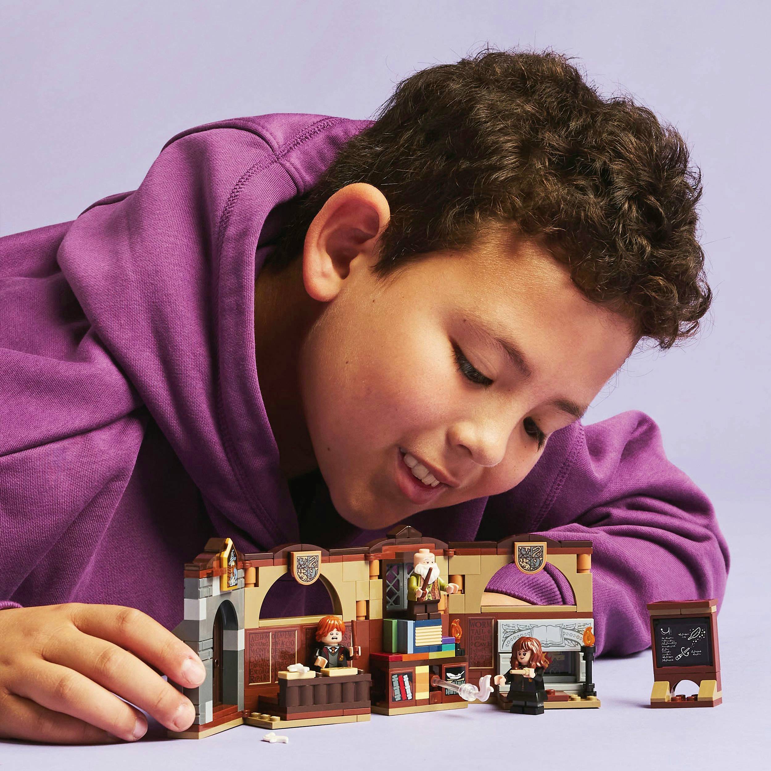 Een kind speelt met Lego-poppetjes aan een tafel. De Lego-scène toont een ruimte met een schoolbord en verschillende figuren. Het kind draagt een paarse hoodie.