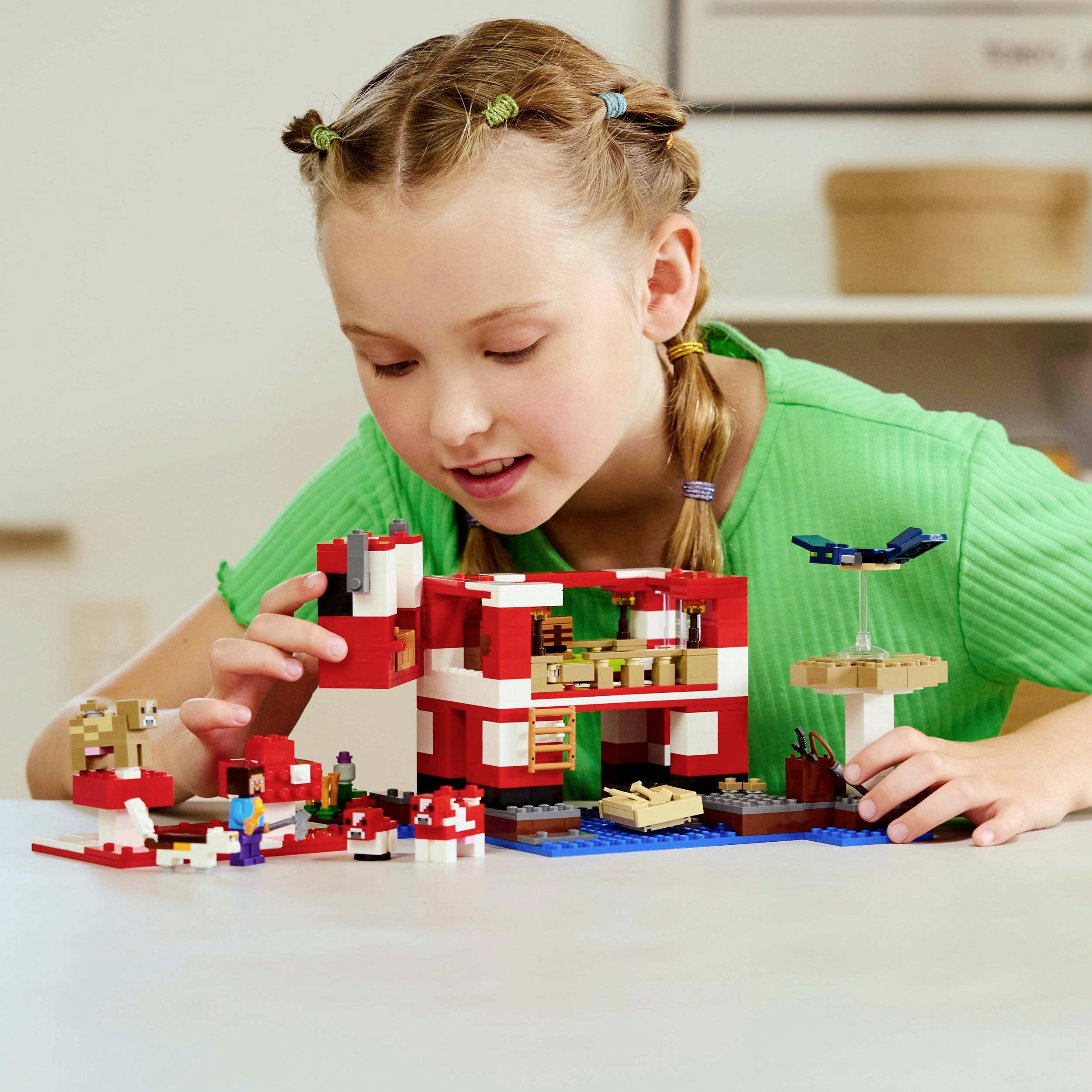 Een meisje bouwt een Lego-huis aan een tafel. Ze draagt een groen bovenste deel en lijkt geconcentreerd en blij.