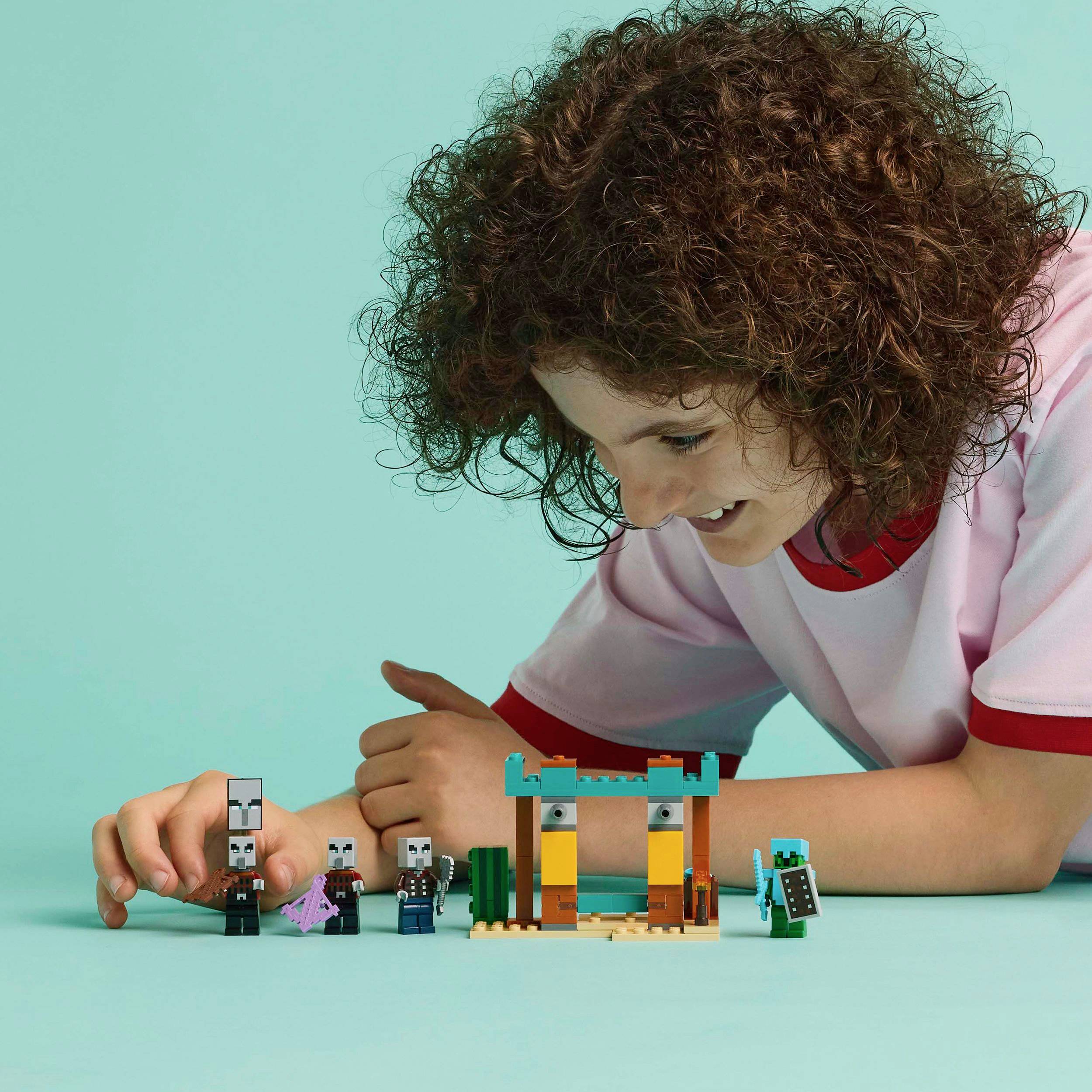 Een kind speelt met blokken en figuren op een tafel. Het draagt een wit shirt met rode randen en glimlacht geconcentreerd.