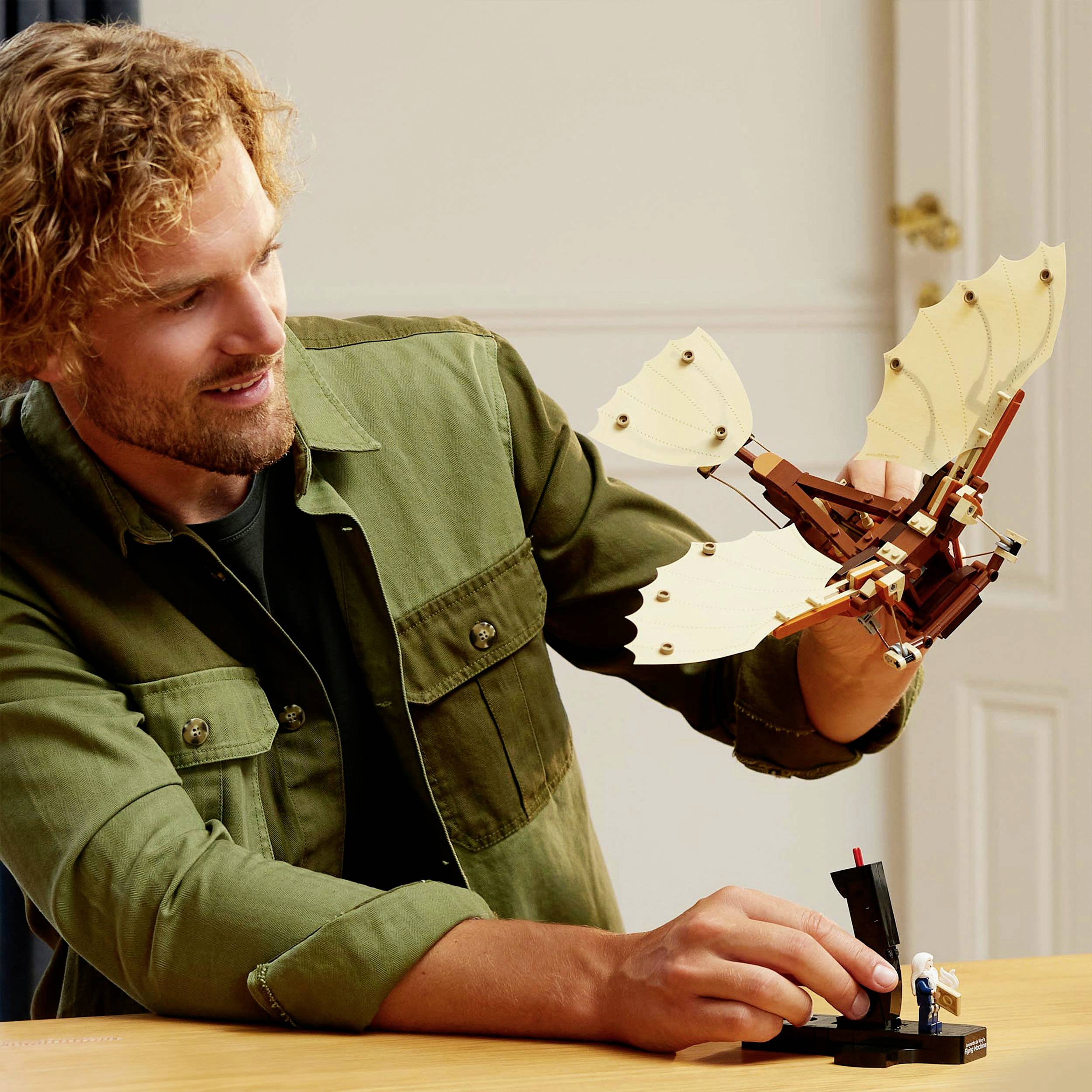 Een persoon bekijkt en houdt een model van een gevleugeld toestel vast. Op de achtergrond is een deur en een houten tafel zichtbaar.