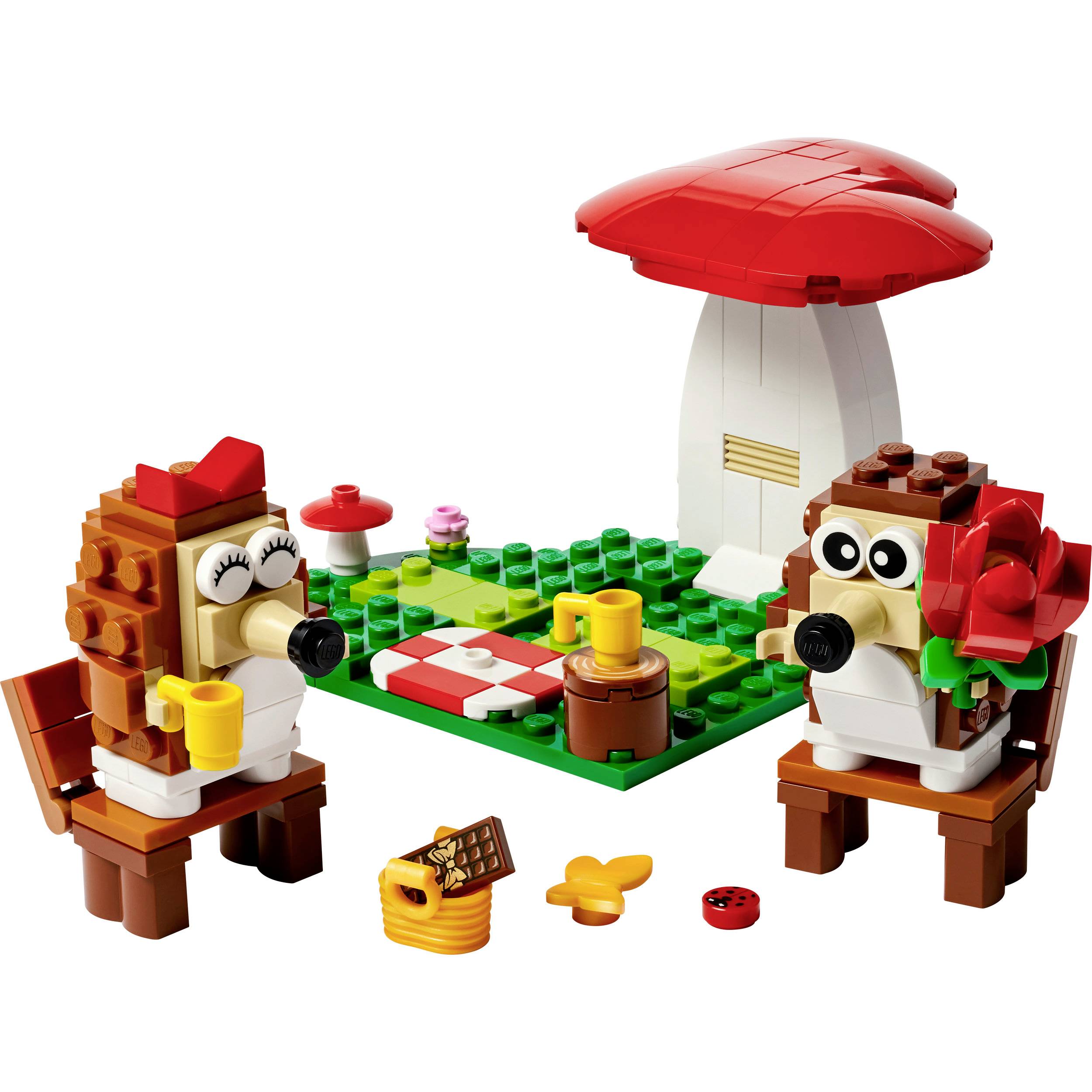 Twee LEGO-egels op een picknick met een paddenstoelhuis op de achtergrond. Eén houdt een bloem, de ander een kopje, op een picknicklaken.