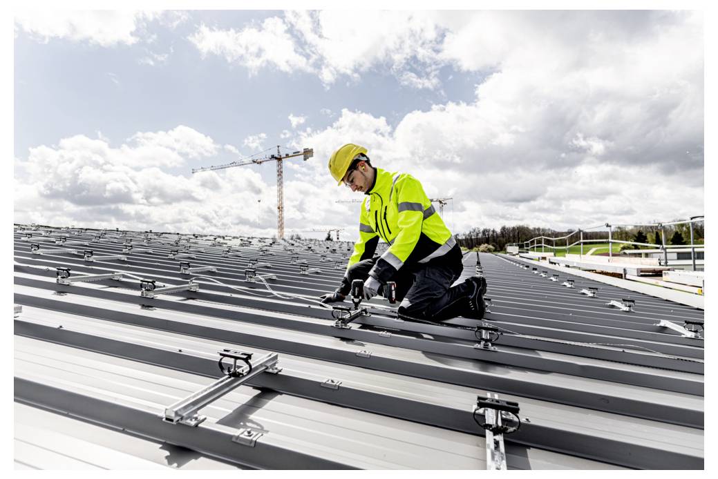 Bouwvakker in veiligheidshelm en fluorescerend veiligheidsvest knielend op een metalen dak, bezig met het bevestigen van zonnepaneel-montagerails onder een bewolkte hemel.