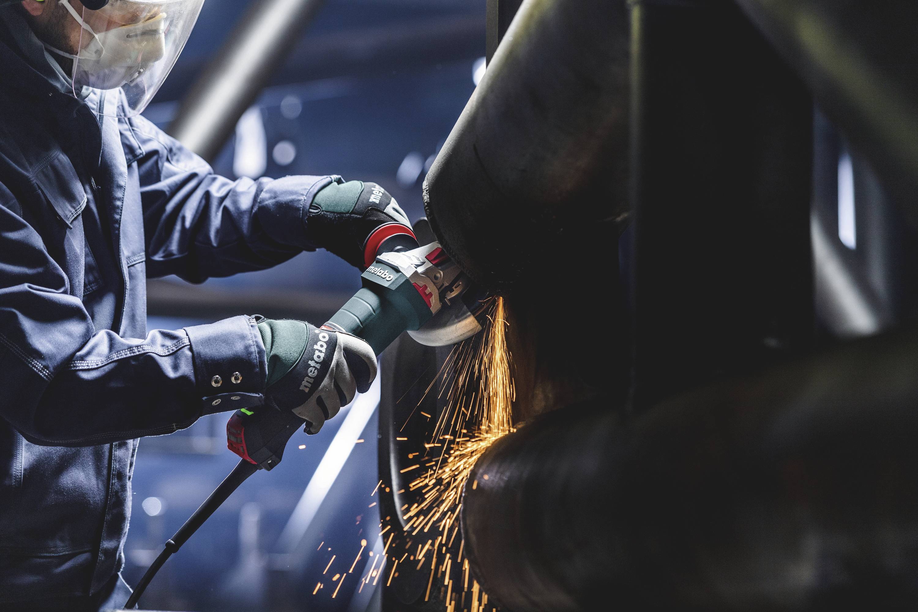 Een persoon draagt beschermende kleding en gebruikt een elektrisch gereedschap om een groot metalen pijp te slijpen. Vonken spatten rond.