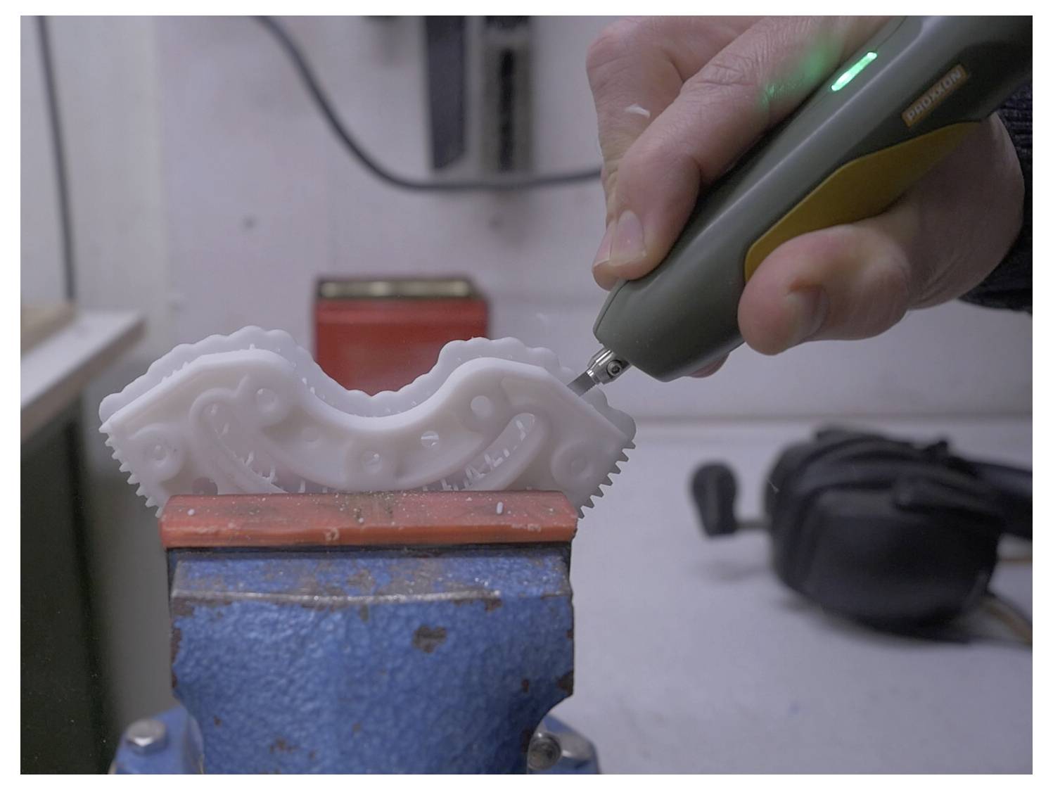 Een hand gebruikt een precisietool om te snijden in een wit kunststof tandwiel dat wordt vastgehouden in een blauwe bankschroef, met de focus op gedetailleerd vakmanschap in een werkplaatsomgeving.