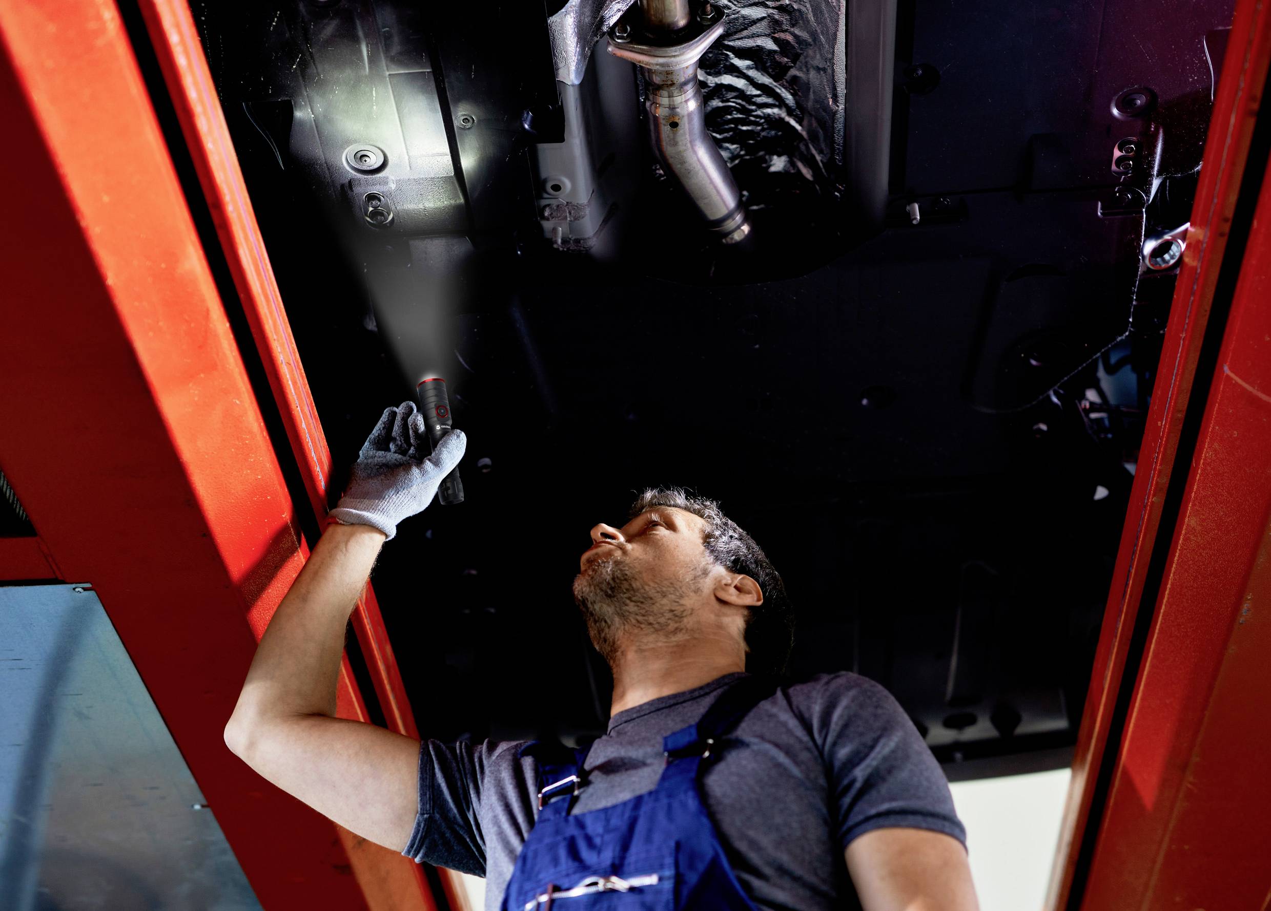 Een monteur in blauwe overall inspecteert de onderkant van een voertuig in een werkplaats, terwijl hij schijnt met een zaklamp.