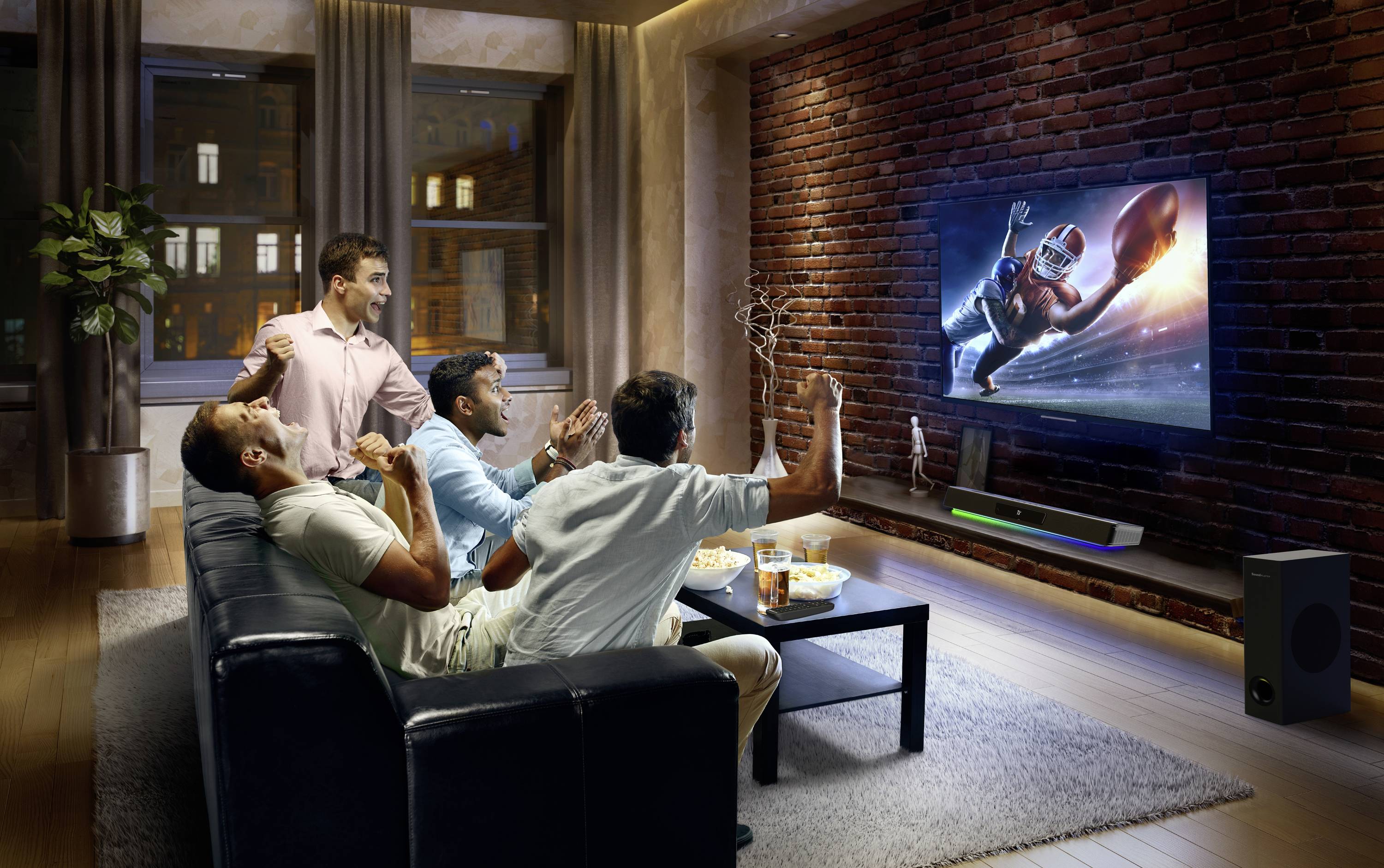 Vier mannen juichen op een bank terwijl ze een American-footballwedstrijd op tv kijken. Snacks staan op een tafel.