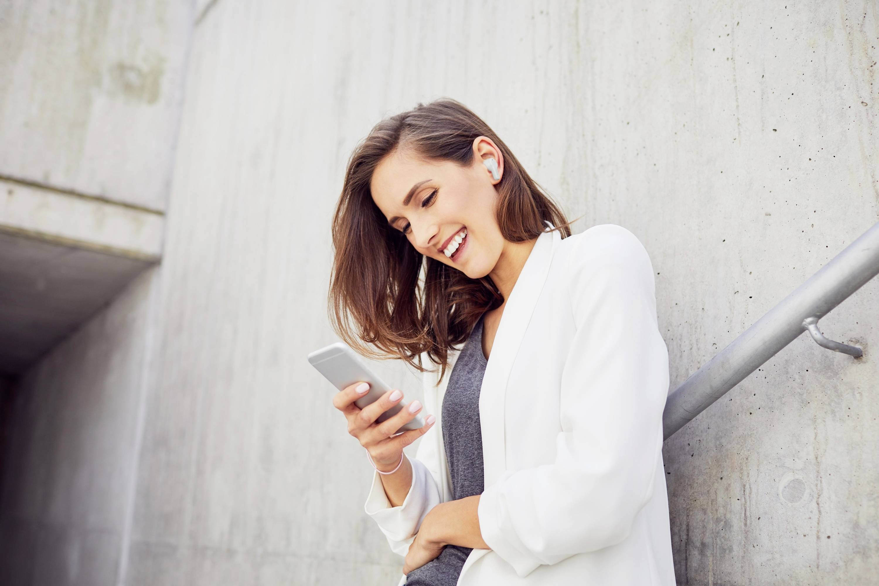 Een vrouw leunt glimlachend tegen een betonnen muur terwijl ze naar haar smartphone kijkt. Ze draagt koptelefoons en een witte jas.