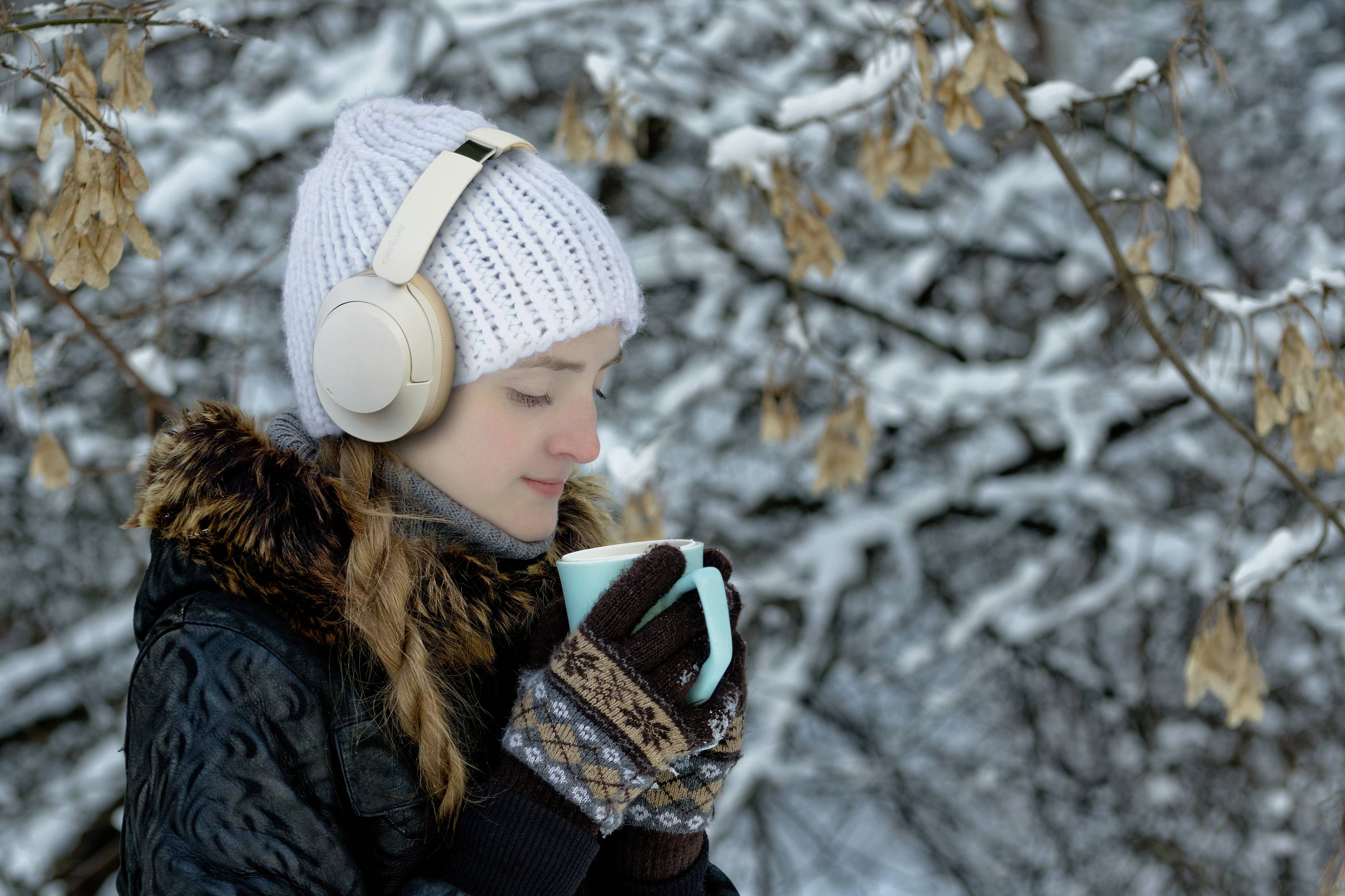 Een persoon in winterkleding geniet van een warme drank buiten. Ze draagt hoofdtelefoons en staat voor een besneeuwde boom.
