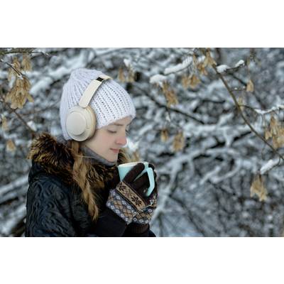 Een persoon in winterkleding geniet van een warme drank buiten. Ze draagt hoofdtelefoons en staat voor een besneeuwde boom.