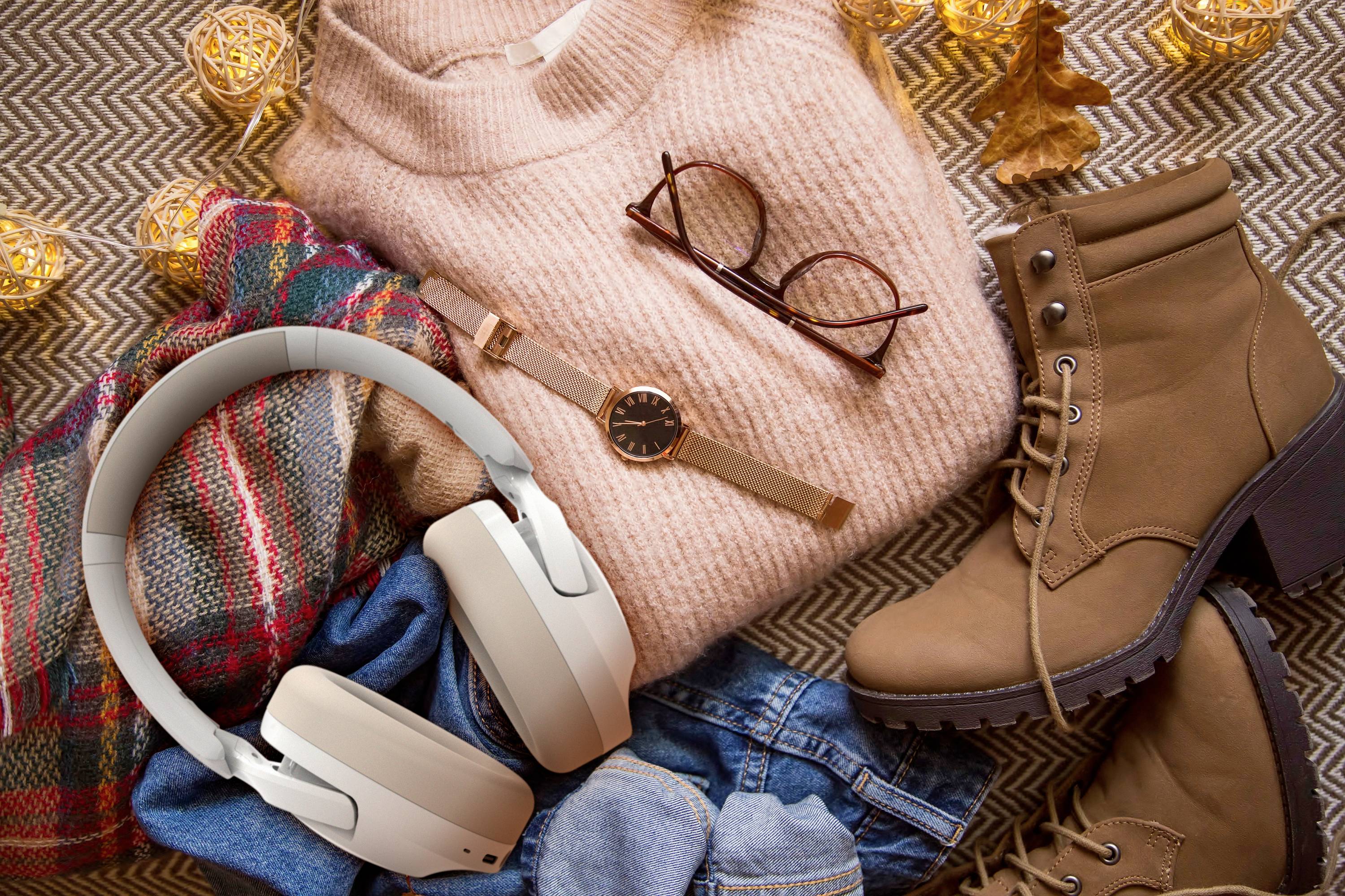 Trui, bril, horloge, koptelefoon, laarzen en sjaal liggen op een deken. Herfstelijke sfeer met warme kleurtinten en gezellige accessoires.