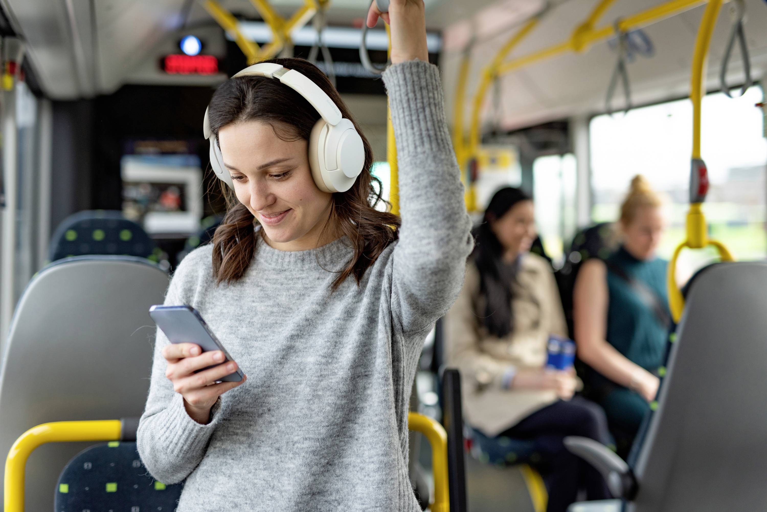 Een vrouw draagt koptelefoons, houdt zich vast in een bus en kijkt glimlachend naar haar smartphone. Andere passagiers zitten op de achtergrond.