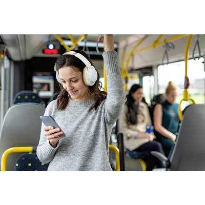 Een vrouw draagt koptelefoons, houdt zich vast in een bus en kijkt glimlachend naar haar smartphone. Andere passagiers zitten op de achtergrond.