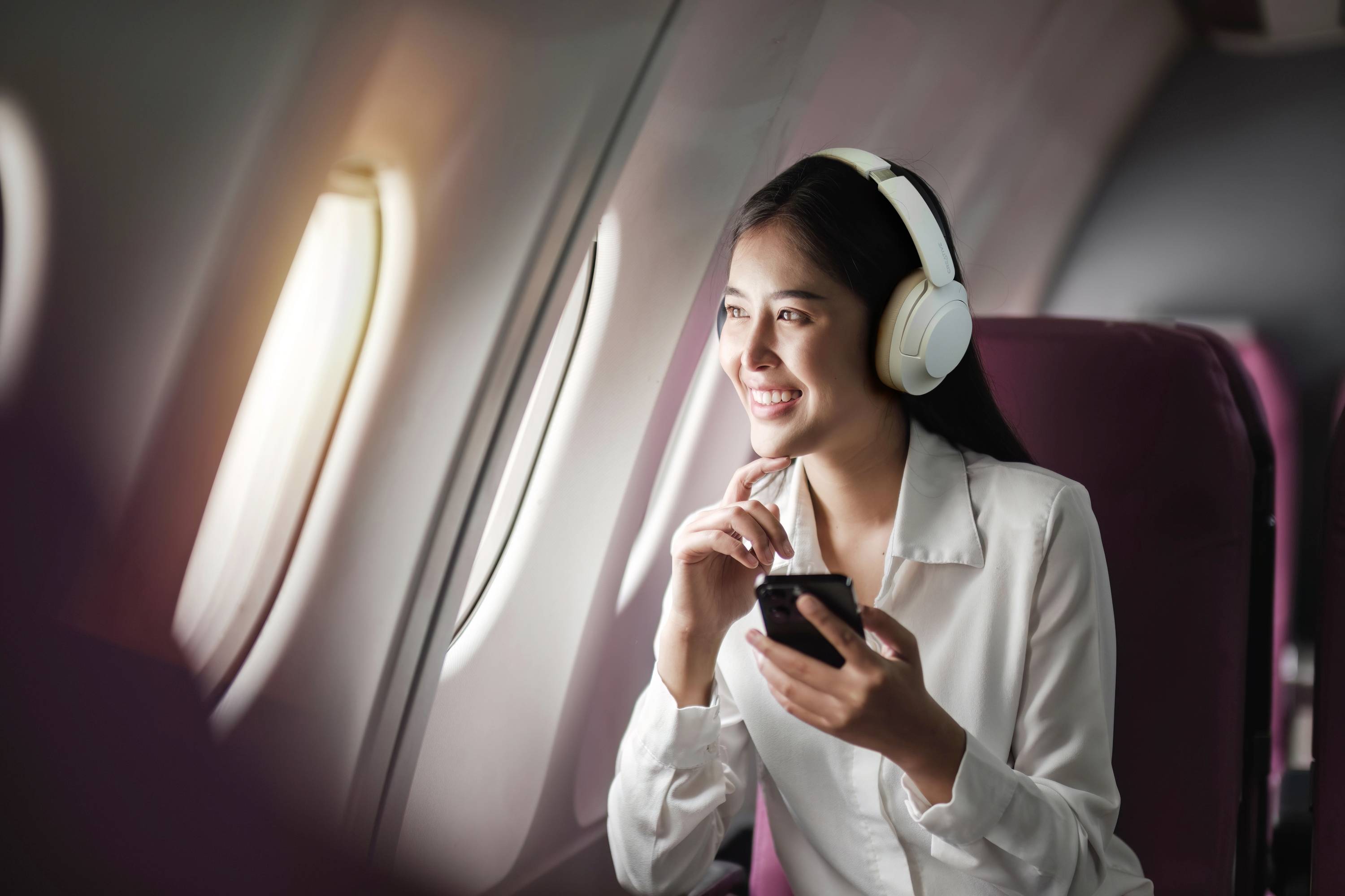 Vrouw met koptelefoon zit in het vliegtuig, houdt een mobiele telefoon vast en kijkt glimlachend uit het raam.