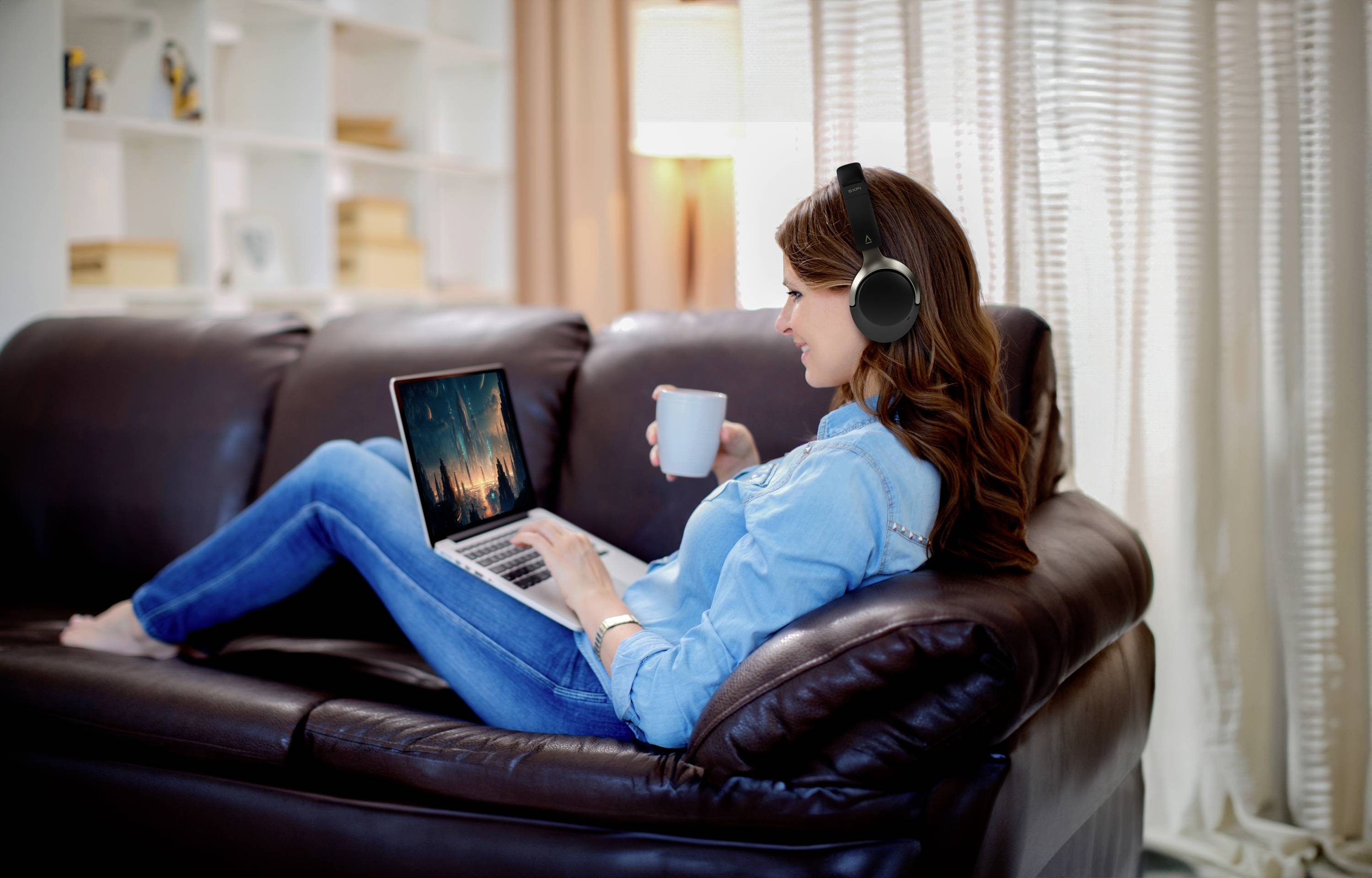 Vrouw zit ontspannen op de bank, draagt koptelefoon, houdt een mok vast en kijkt naar het laptop-scherm. Gezellige, moderne woonkamer.