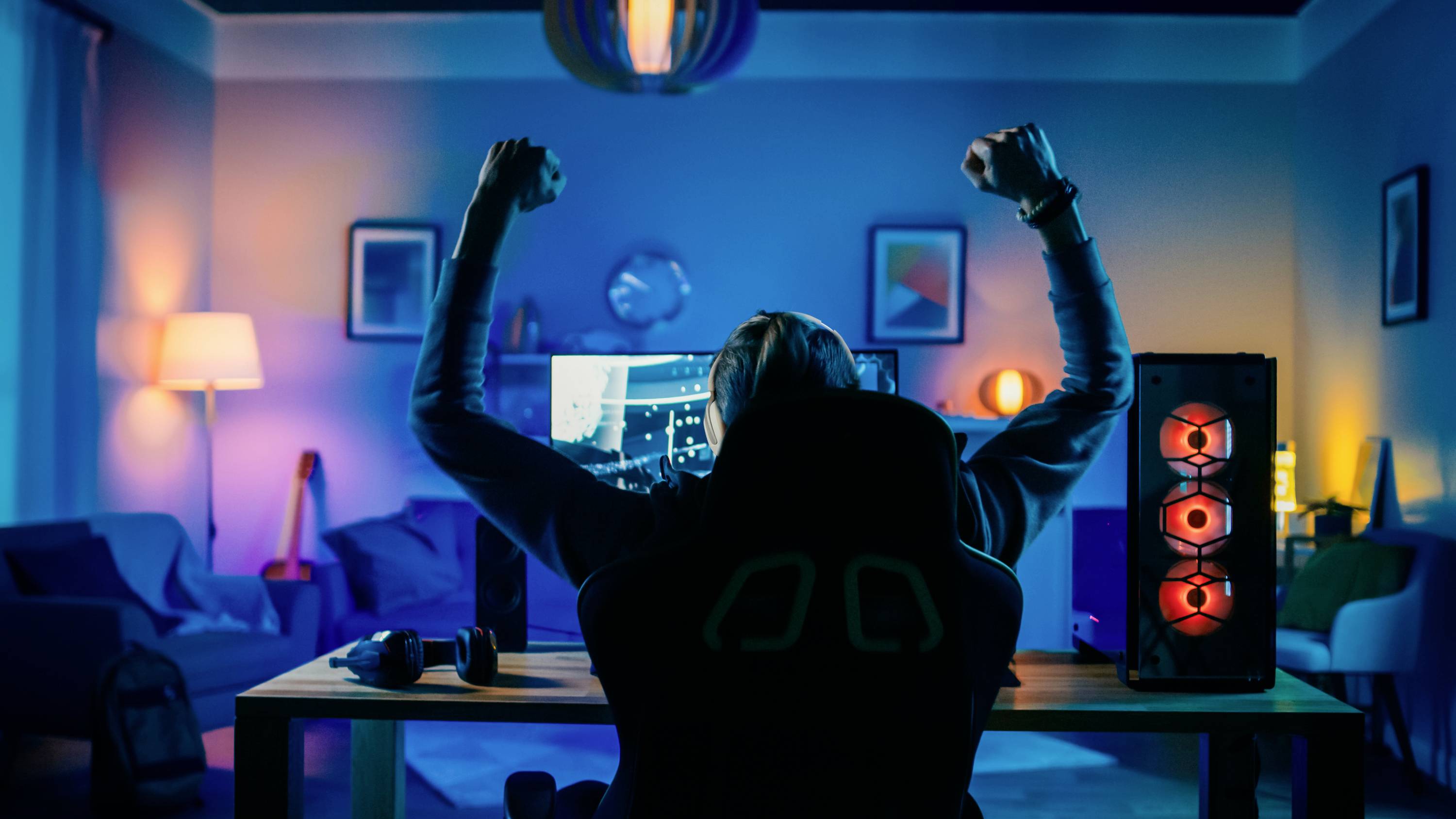 Een persoon zit aan een bureau en steekt juichend de armen in de lucht voor een scherm met een videospel. De ruimte is kleurrijk verlicht.