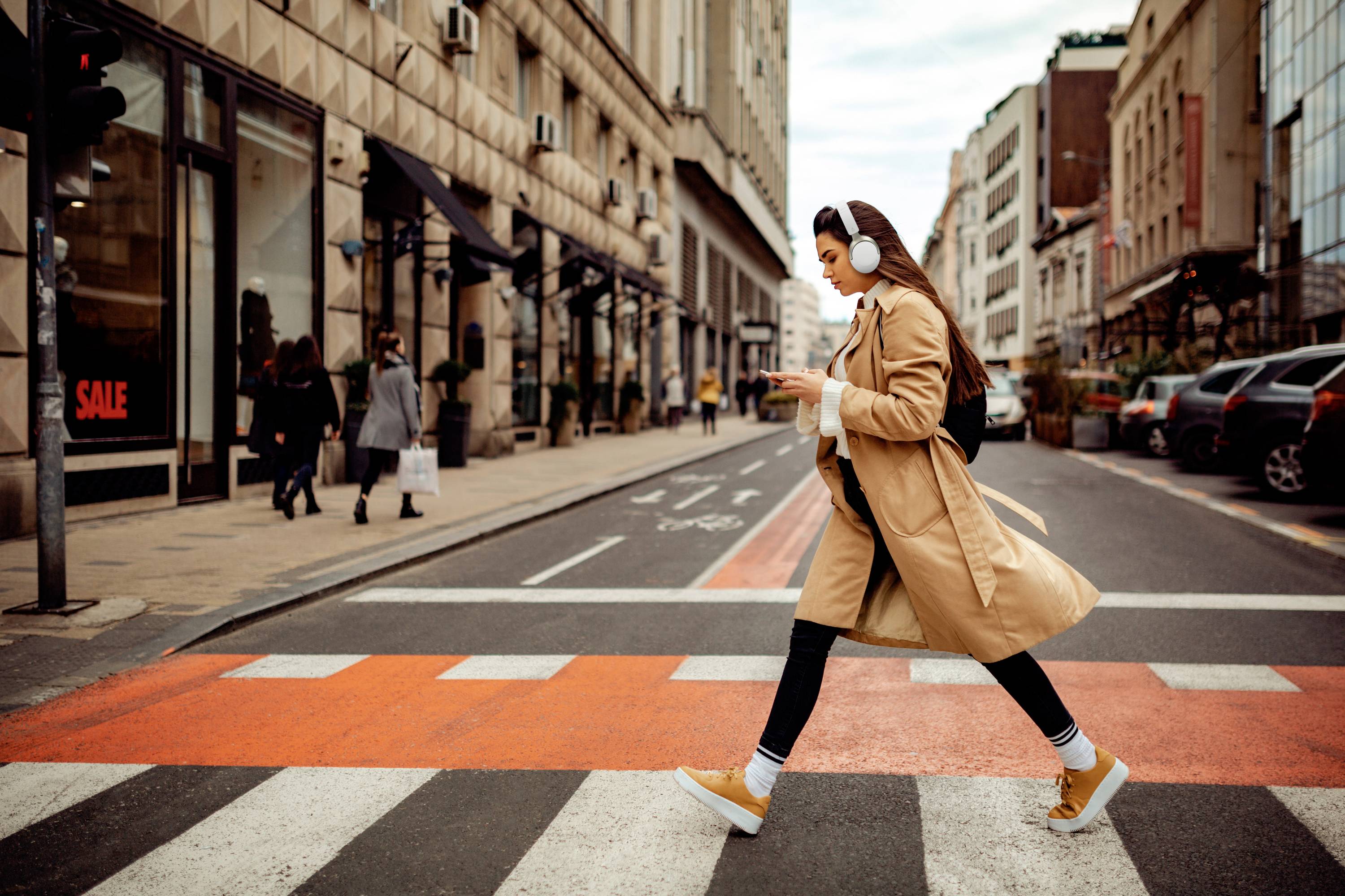 Een vrouw steekt overdag een zebrapad over in een stadsstraat. Ze draagt koptelefoons en kijkt naar haar mobiele telefoon.