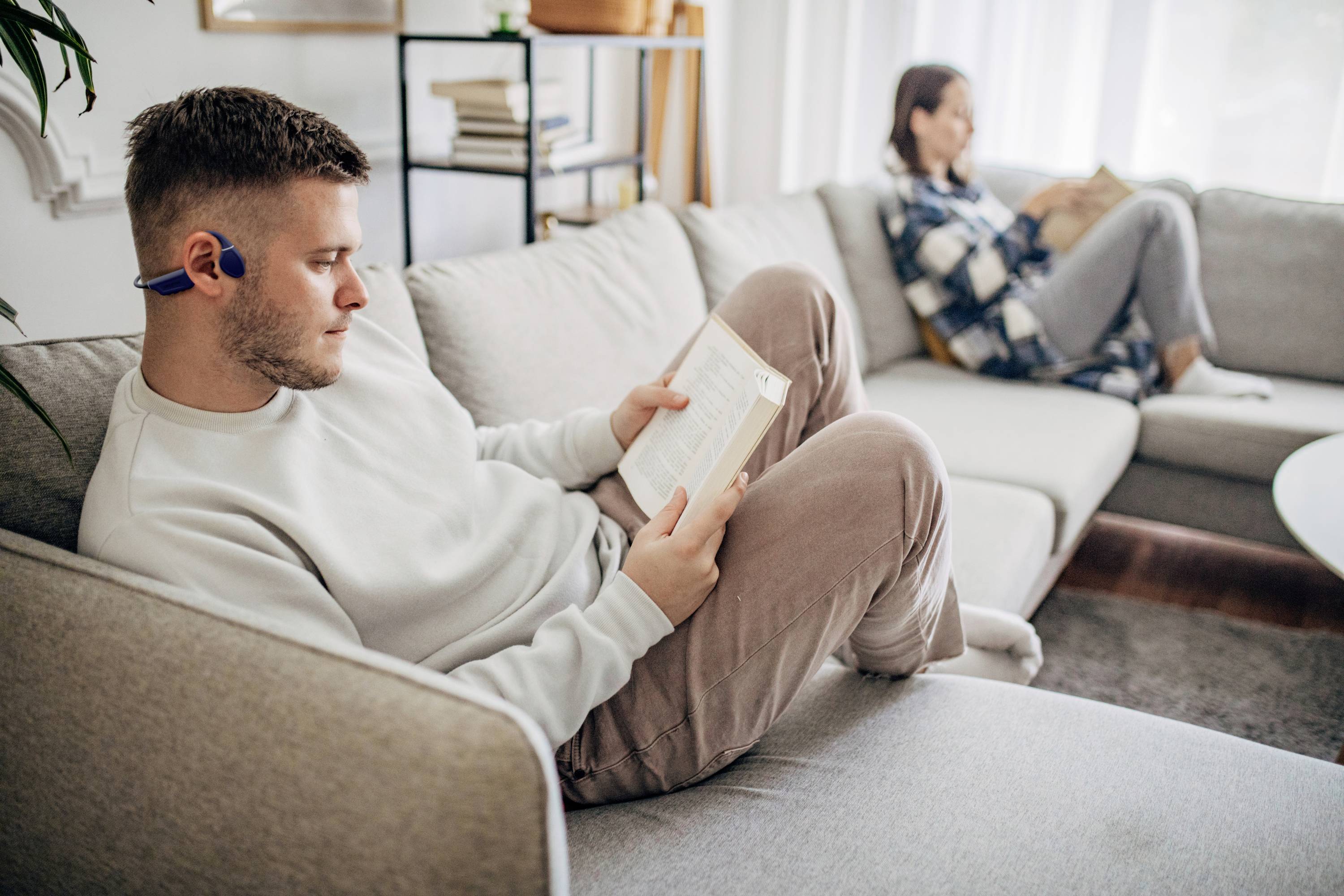 Een persoon leest een boek op een bank en draagt koptelefoons, terwijl een andere persoon op de achtergrond ook aan het lezen is.