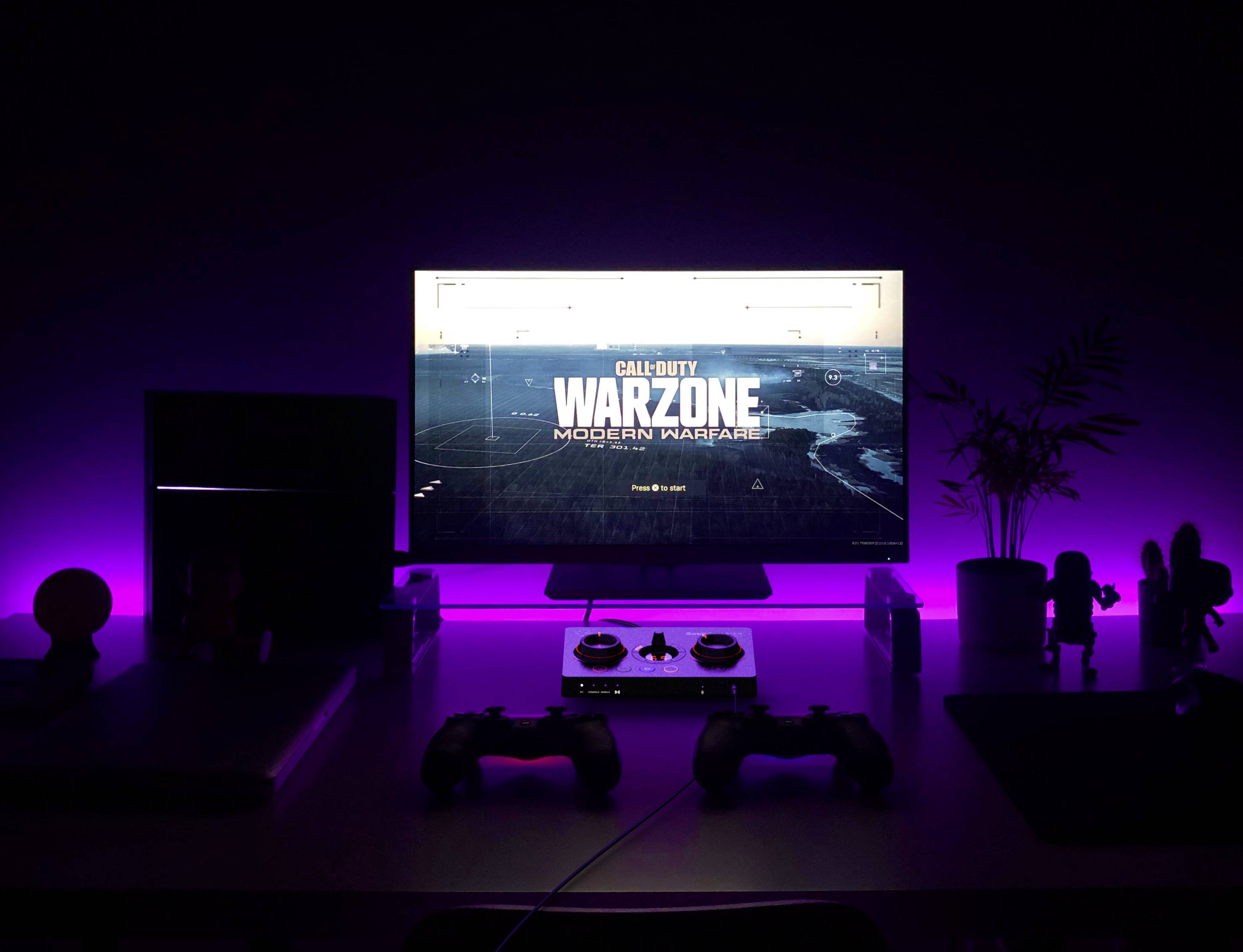 Een gaming-setup met paars achtergrondlicht toont een scherm met het spel 'Call of Duty Warzone'. Voor de monitor liggen controllers.
