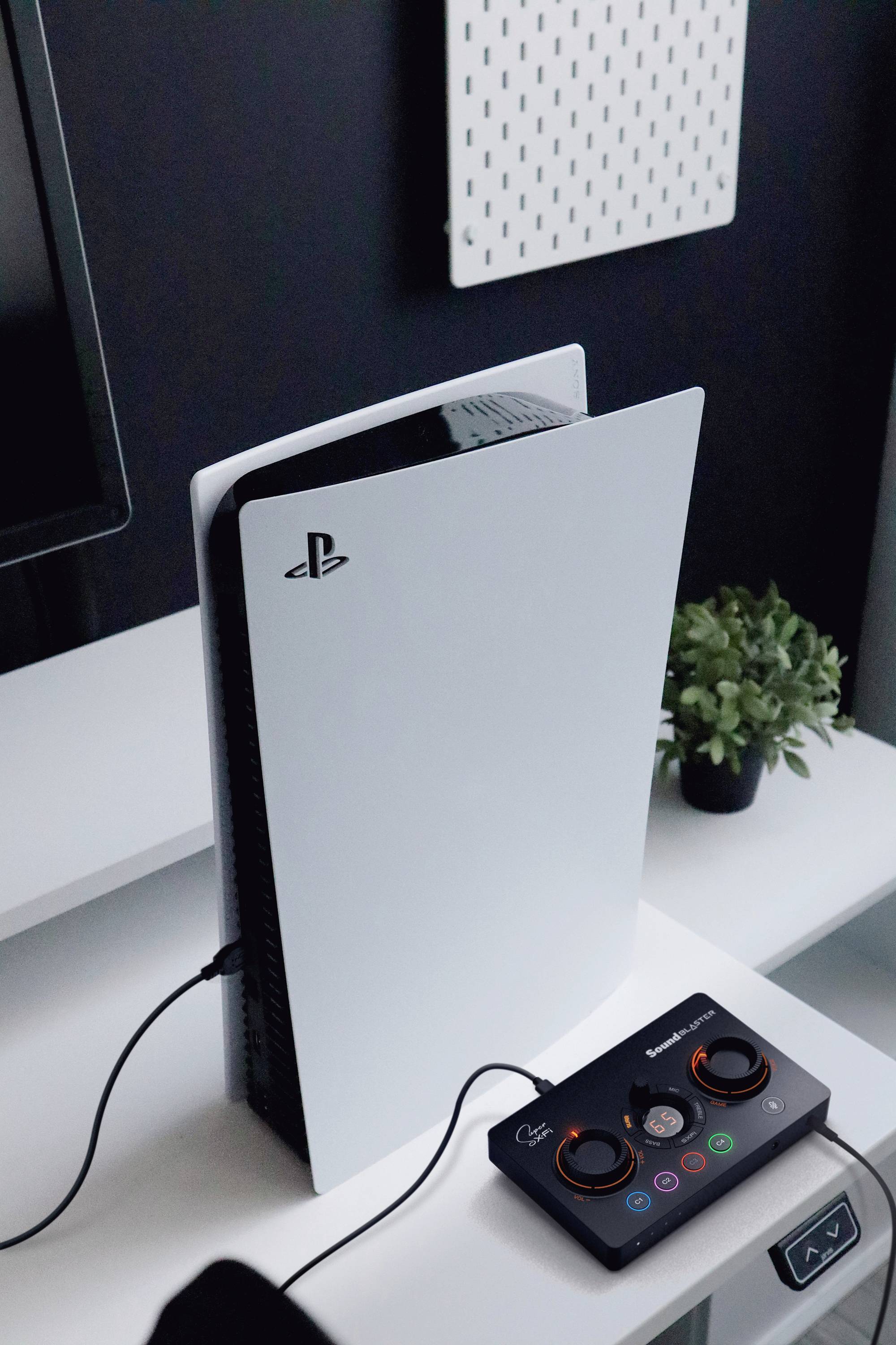 PlayStation 5-console op een tafel, aangesloten op een zwart accessoire-apparaat met meerdere verlichte knoppen. Planten op de achtergrond.