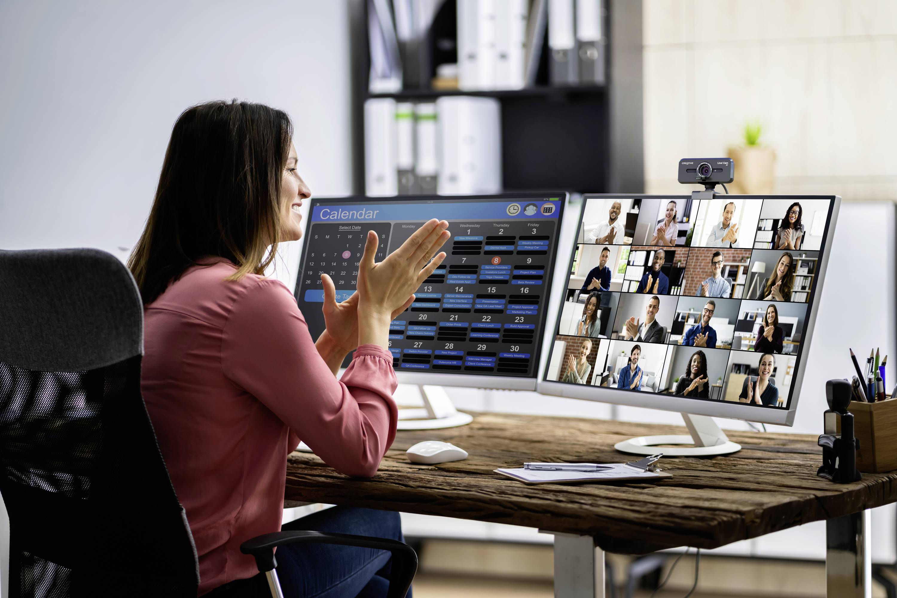 Een vrouw zit aan een bureau en neemt deel aan een videoconferentie met meerdere deelnemers op twee monitoren.