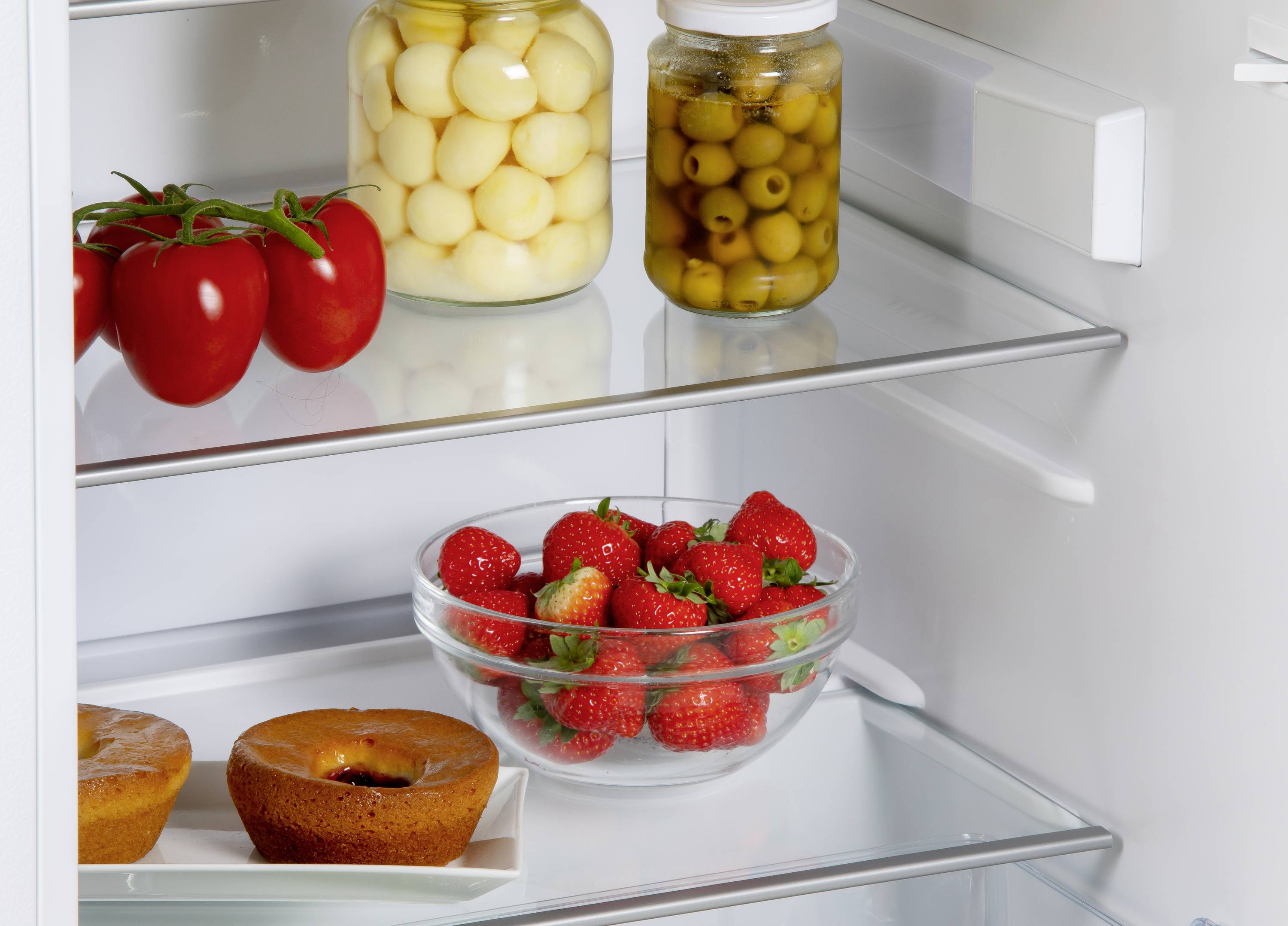 Tomaten, een schaaltje aardbeien, ingelegde uitjes, olijven en twee donuts bevinden zich op verschillende schappen in de open koelkast.