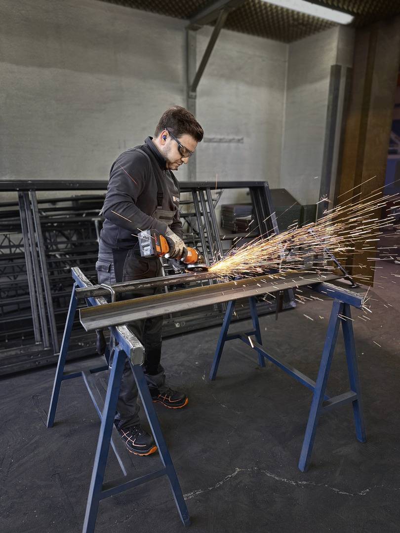 Een persoon slijpt metalen buizen met een haakse slijper. Vonken spatten rond. Veiligheidsuitrusting zoals een veiligheidsbril en handschoenen worden gedragen.