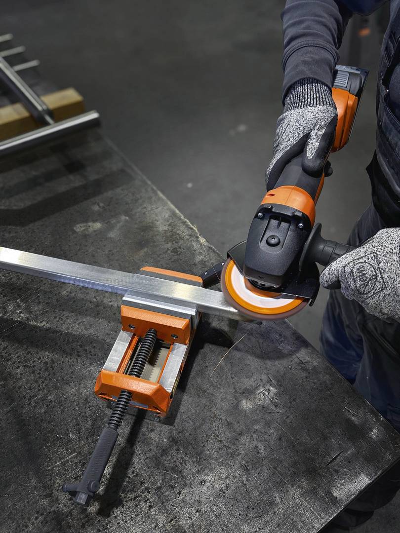 Een persoon slijpt metalen randen met een haakse slijper, waarbij het metalen werkstuk is vastgeklemd in een bankschroef, en draagt werkhandschoenen.