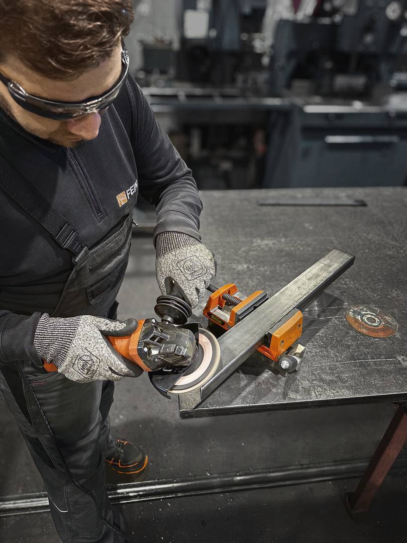 Een persoon bewerkt met een haakse slijper een metalen stuk dat met een bankschroef op een werkbank is vastgezet.