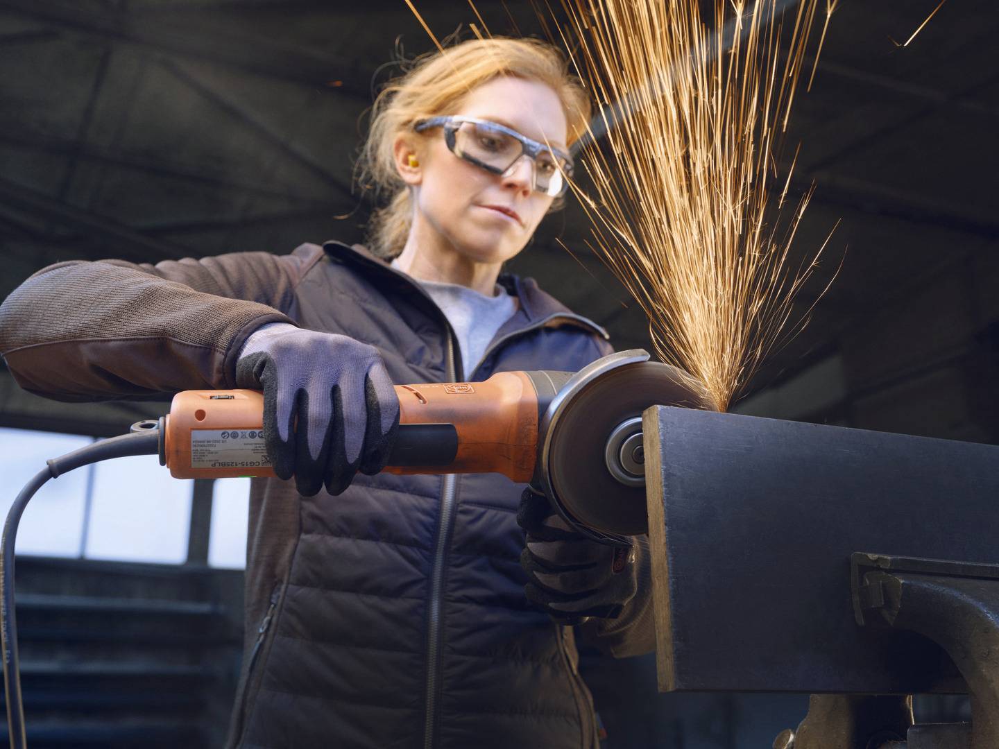 Een persoon slijpt metaal met een haakse slijper, vonken spatten. Hij draagt een veiligheidsbril en handschoenen in een werkplaats.