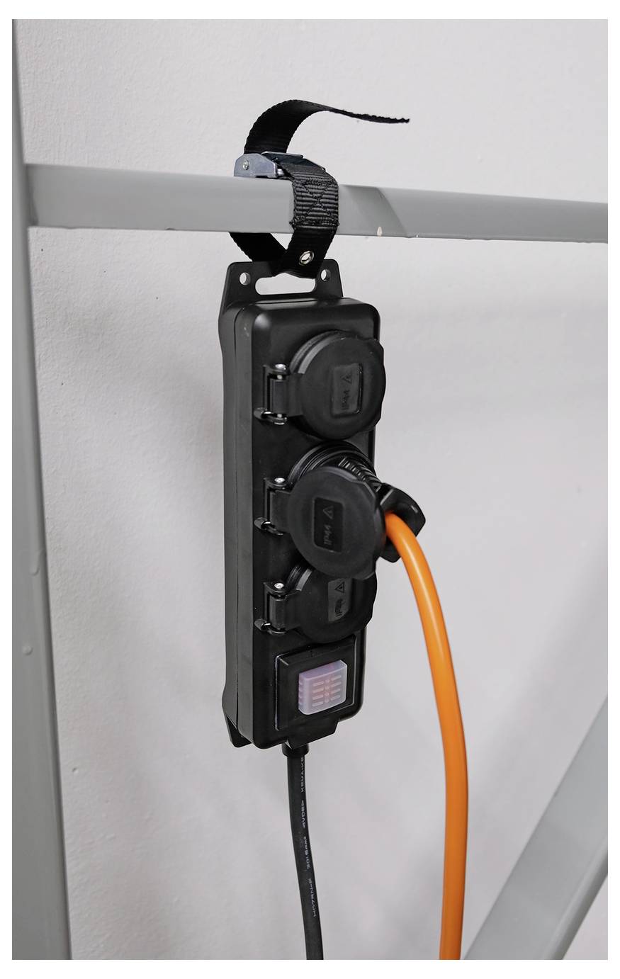 Stekkerdoos hangend aan een metalen rail, met een zwart snoer en oranje kabel erop aangesloten tegen een kale muur.