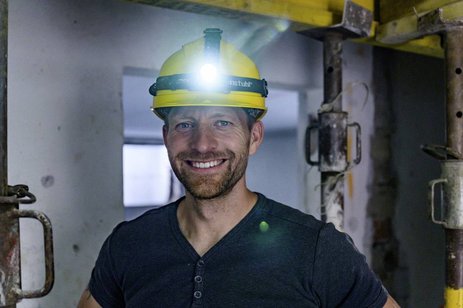 Een lachende man met een gele bouwhelm en hoofdlamp staat in een casco-ruimte. Hij draagt een donker T-shirt.