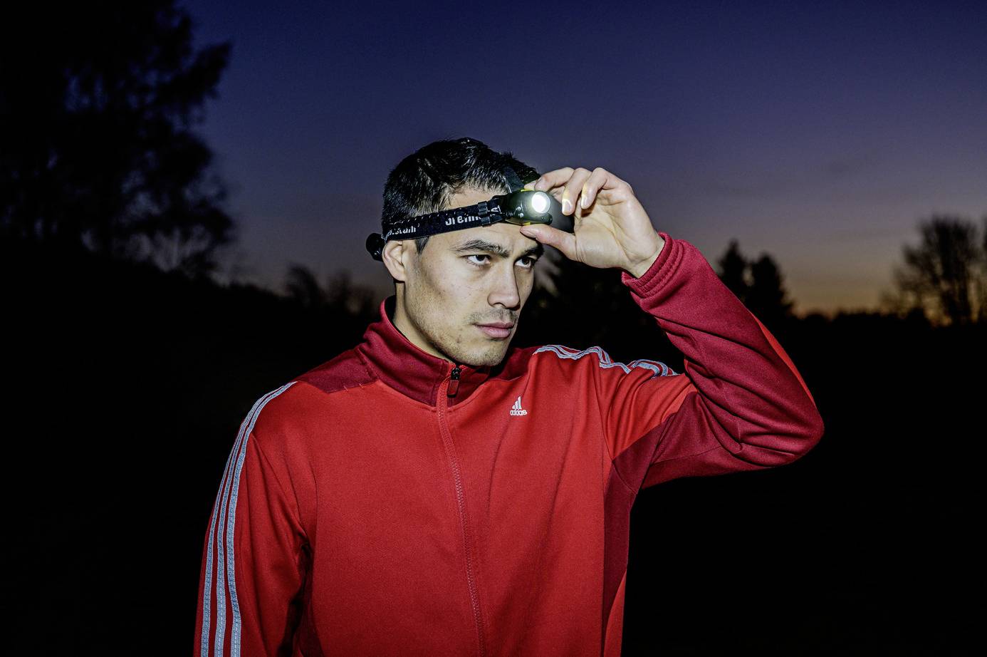 Een man in een rode trainingsbroek staat buiten bij schemering. Hij draagt een hoofdlamp die schijnt en kijkt geconcentreerd in de verte.