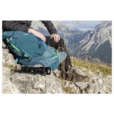 Wandelaar die rust op een rotsachtig bergplateau met een tealblauw rugzak, laarzen en een cameralens ervan; alpine pieken op de achtergrond.