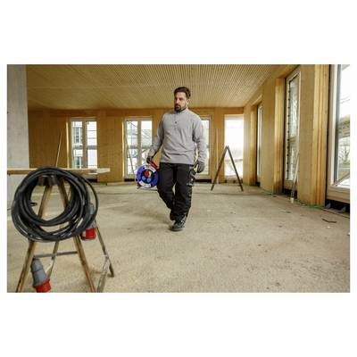 Een man met een kabelspoel loopt door een onafgewerkte kamer tijdens de bouw, met blootgestelde houten wanden, ramen en gereedschap op de grond.