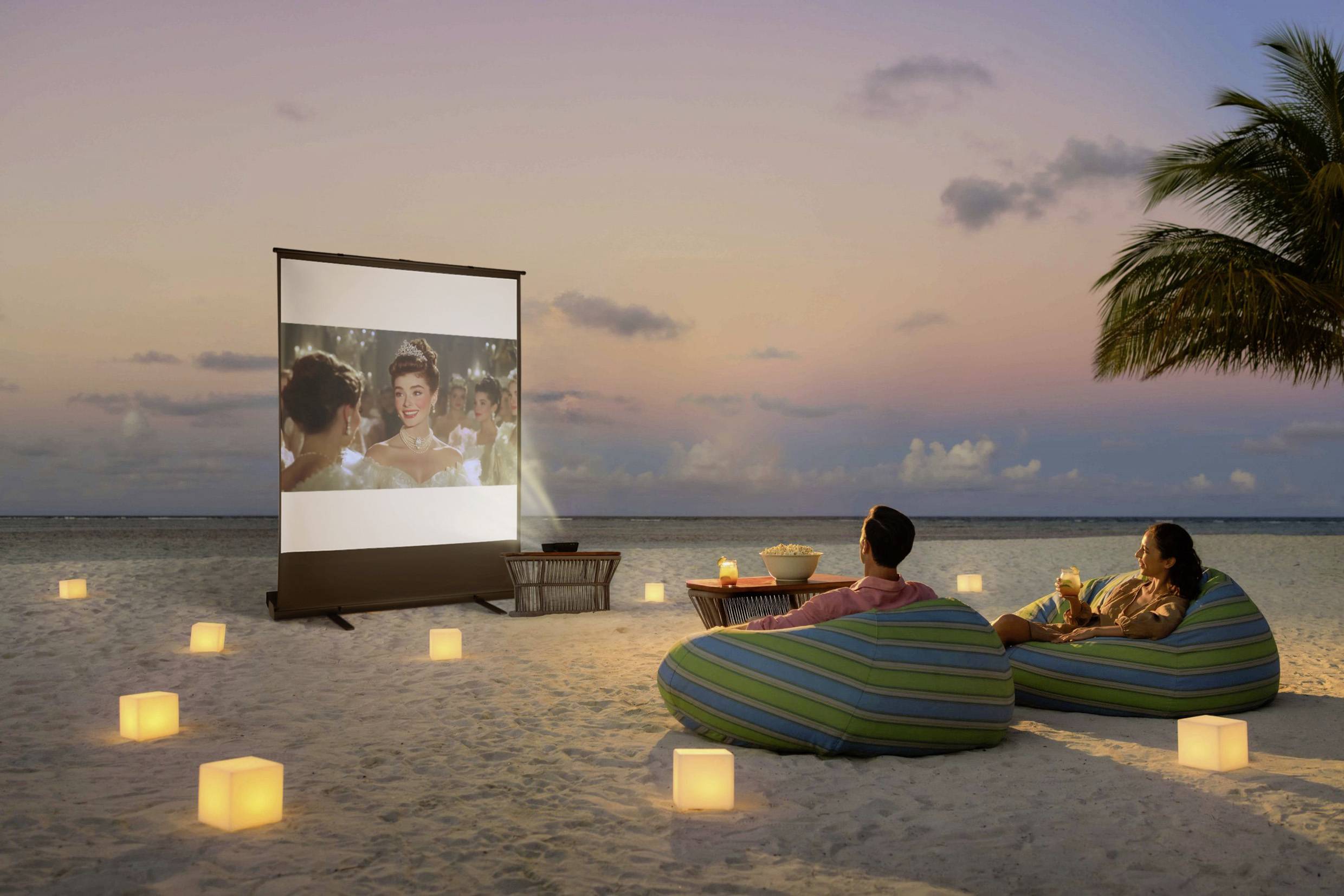 Een stel zit op zitzakken aan het strand en kijkt naar een film op een scherm bij zonsondergang, omringd door lantaarns.