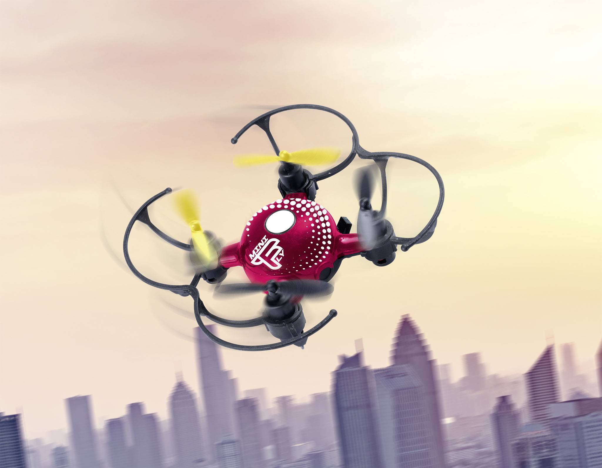 Een rood-zwarte drone met gele propellers vliegt voor een stadslandschap bij zonsondergang.