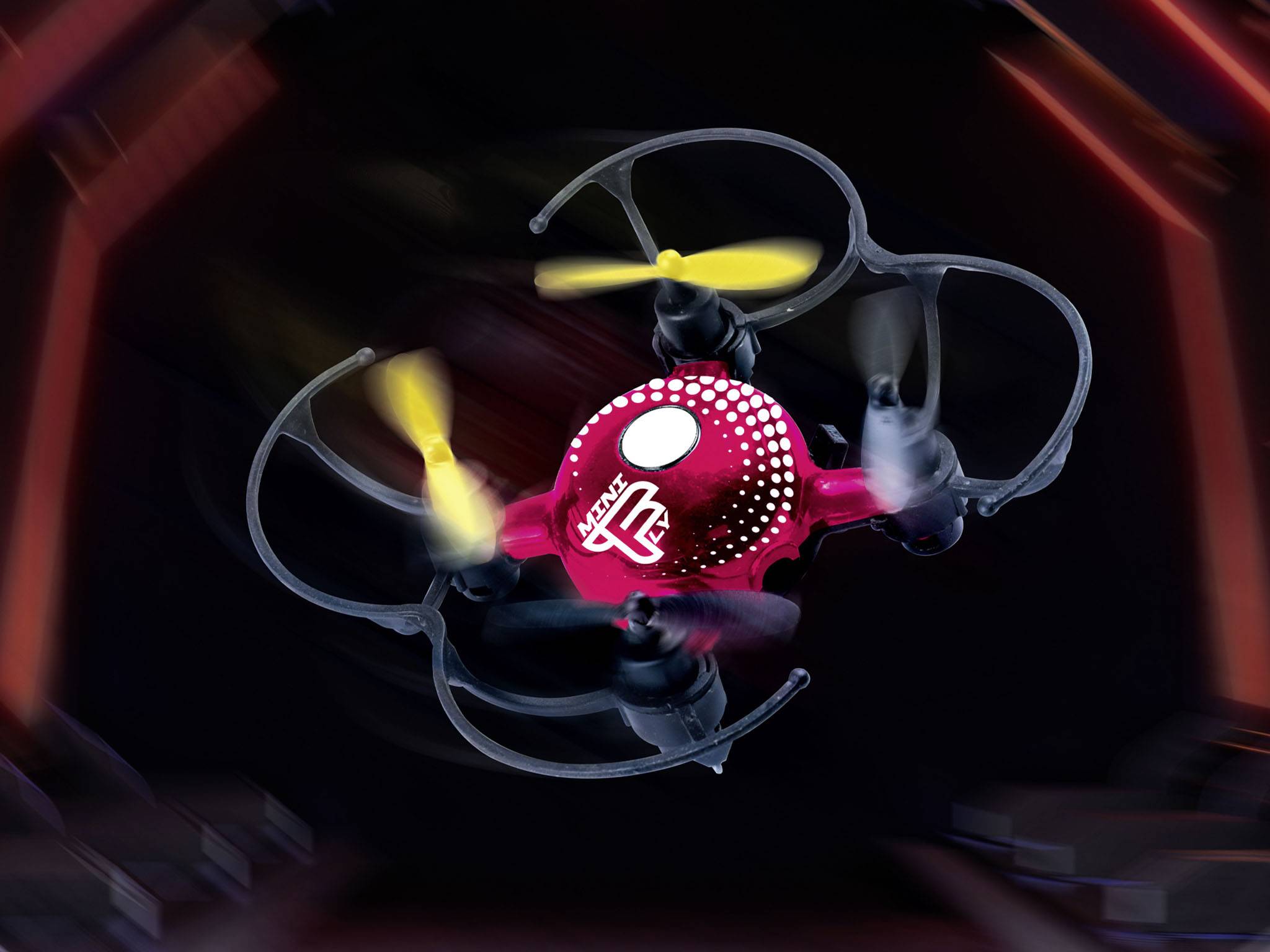 Een rode drone met gele propellers zweeft in de lucht. De achtergrond is wazig en donker, waardoor de drone opvalt.