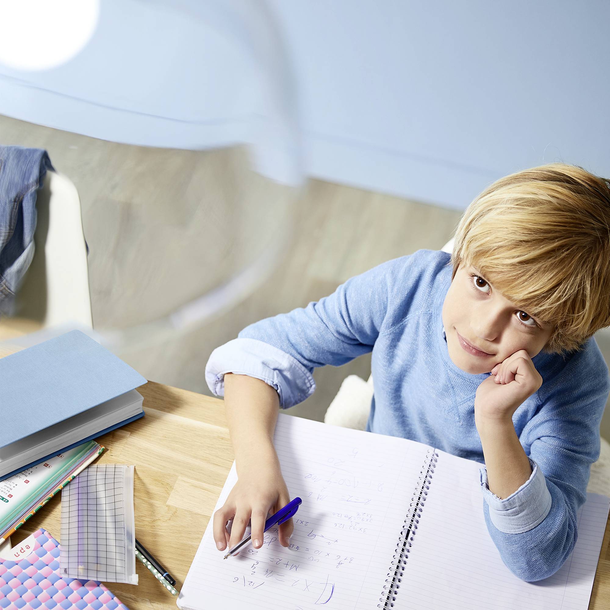 Een jongen met blond haar zit aan een bureau en kijkt nadenkend terwijl hij in een schrift schrijft. Boeken en schrijfmaterialen liggen op tafel.