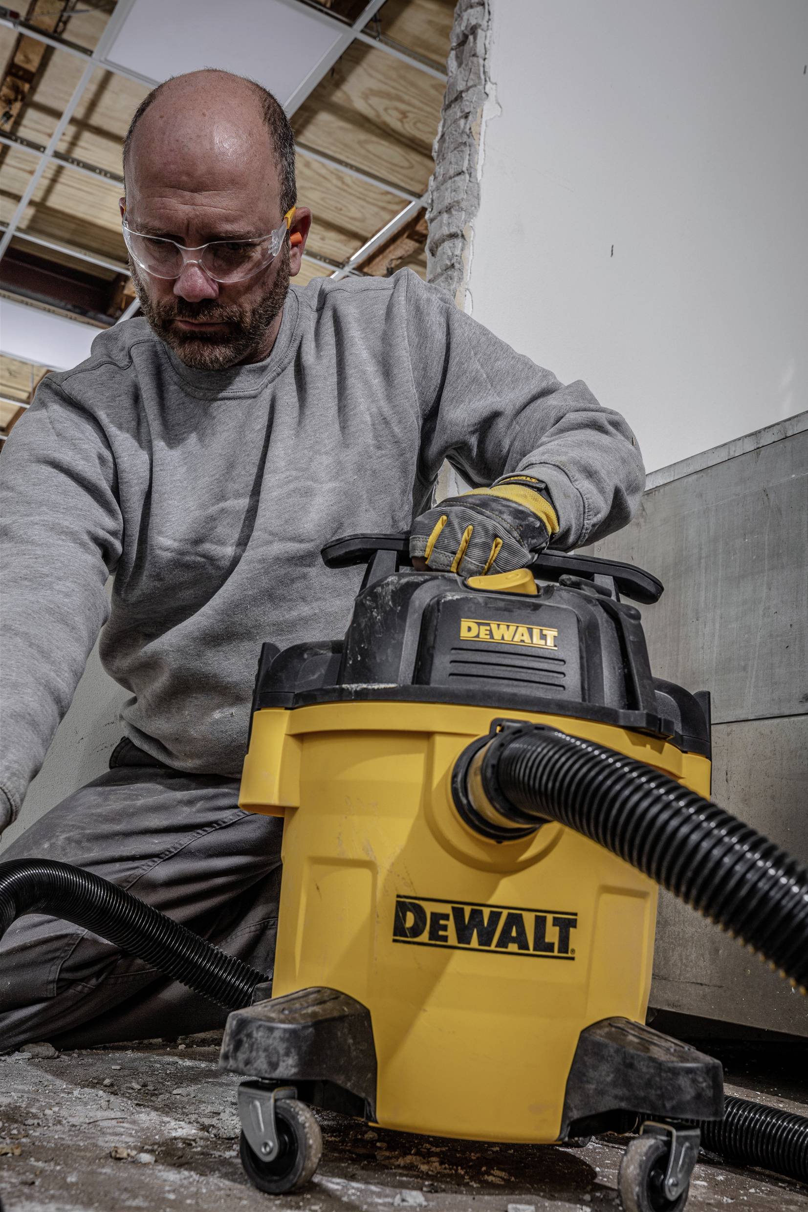 Een persoon reinigt een bouwplaats met een gele nat-/droogzuiger van DeWalt. Hij draagt werkkleding en handschoenen.