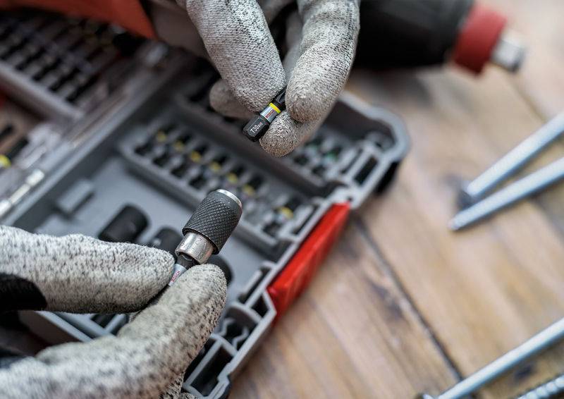 Close-up van handen in werkhandschoenen die bits uit een gereedschapskist vasthouden. Op de achtergrond is een schroevendraaier zichtbaar.