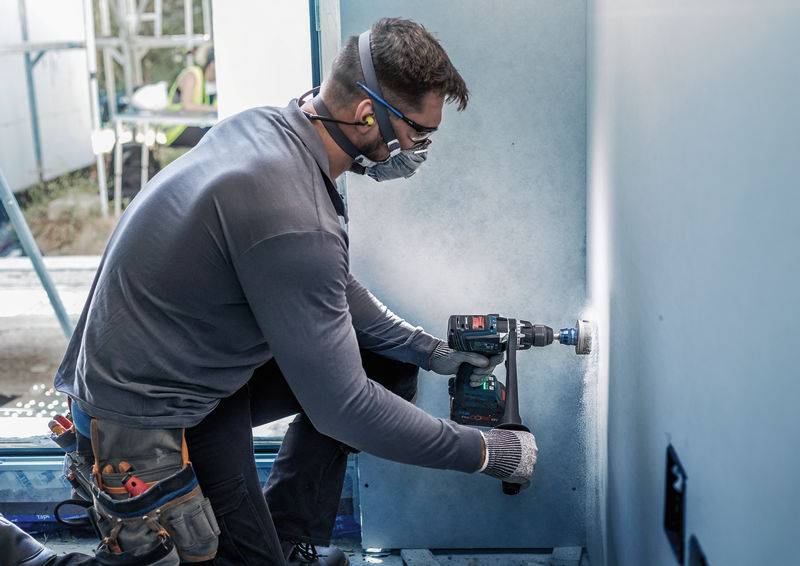 Een man boort met een boormachine in een muur, draagt een veiligheidsbril en een adembeschermingsmasker. Bouwplaatsomgeving op de achtergrond.