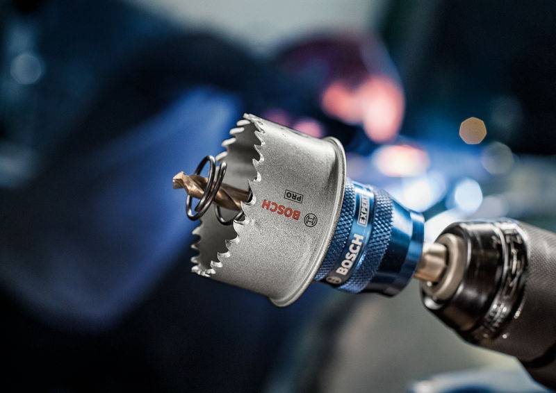 'Close-up van een Bosch-boor met gatenzaagopzetstuk, scherp in beeld vooraan. Onscherpe achtergrond toont een persoon aan het werk.'