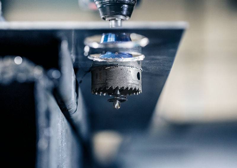 Een metalen boor doorboort een metalen plaat, waarbij metaalspanen zichtbaar zijn. De scène toont een productie- of fabricageproces.