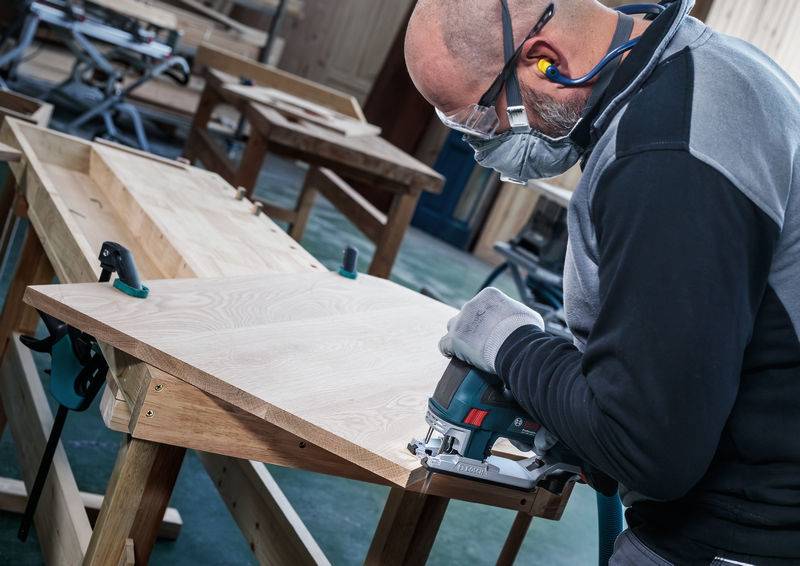 Een man met veiligheidsbril en adembescherming zaagt hout recht met een decoupeerzaag aan een werkbank. Op de achtergrond zijn nog meer werkbanken te zien.