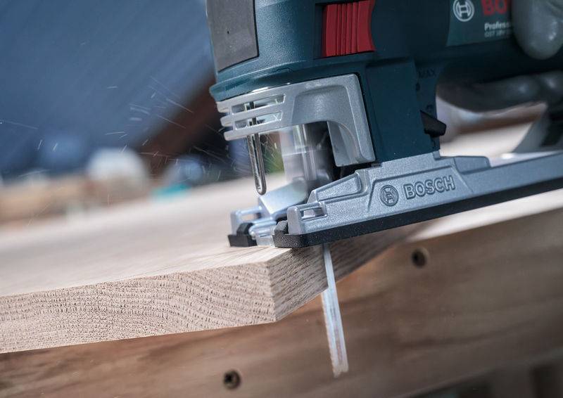 Een elektrische decoupeerzaag snijdt precies door een houten plaat, produceert zaagsel; merk 'Bosch' zichtbaar.