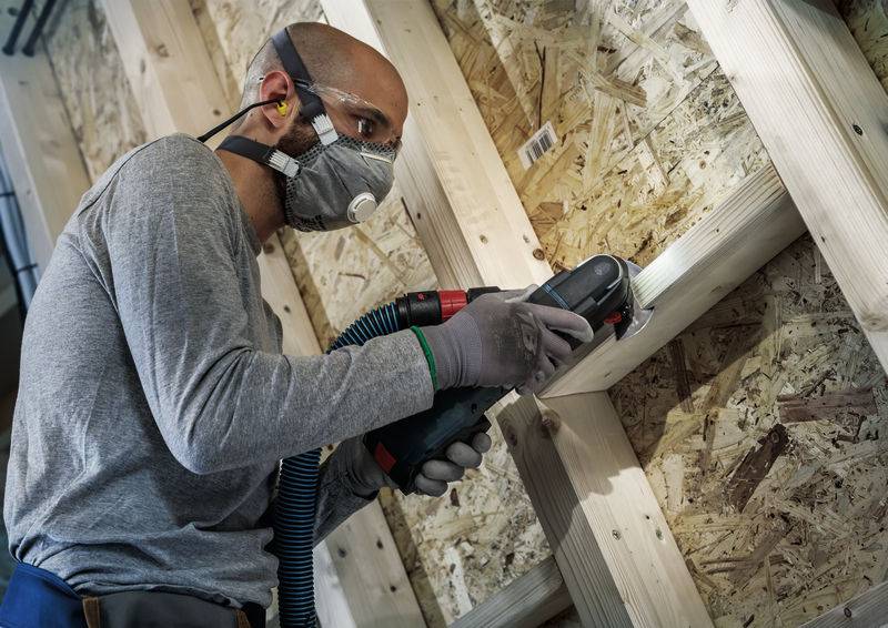 Een ambachtsman slijpt houten balken met een elektrische slijpmachine in een werkplaats. Hij draagt een veiligheidsbril, mondmasker en gehoorbescherming.