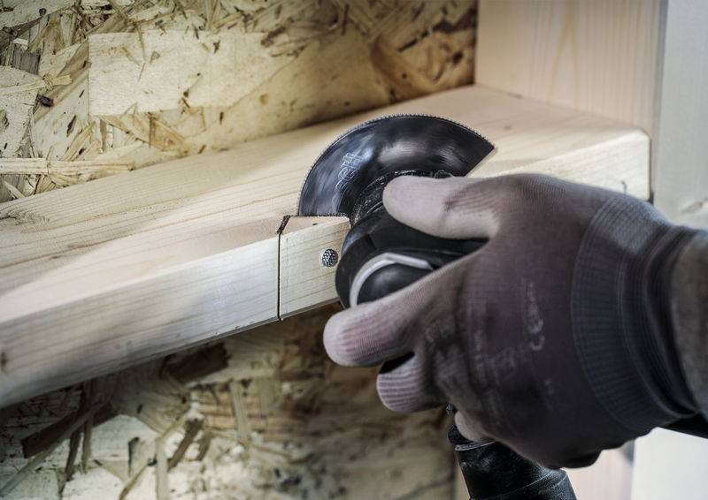 Hand met handschoen houdt een elektrische zaag vast, zaagt door een houten plank voor een spaanplaatmuur.