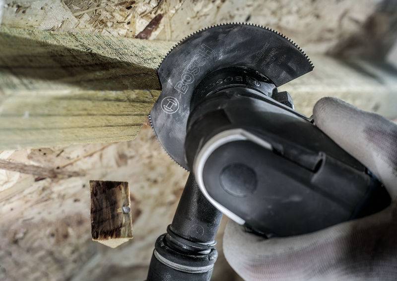 Een hand houdt een elektrisch multifunctioneel gereedschap vast dat een houten plank doorsnijdt. Een stuk hout valt af.