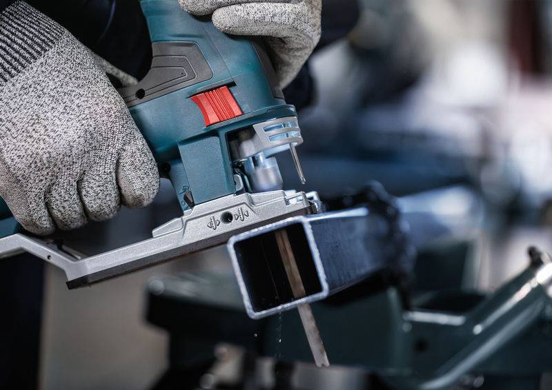 Hand houdt decoupeerzaag, snijdt metalen buis op werkbank. Beschermende kleding draagt bij aan veiligheid.